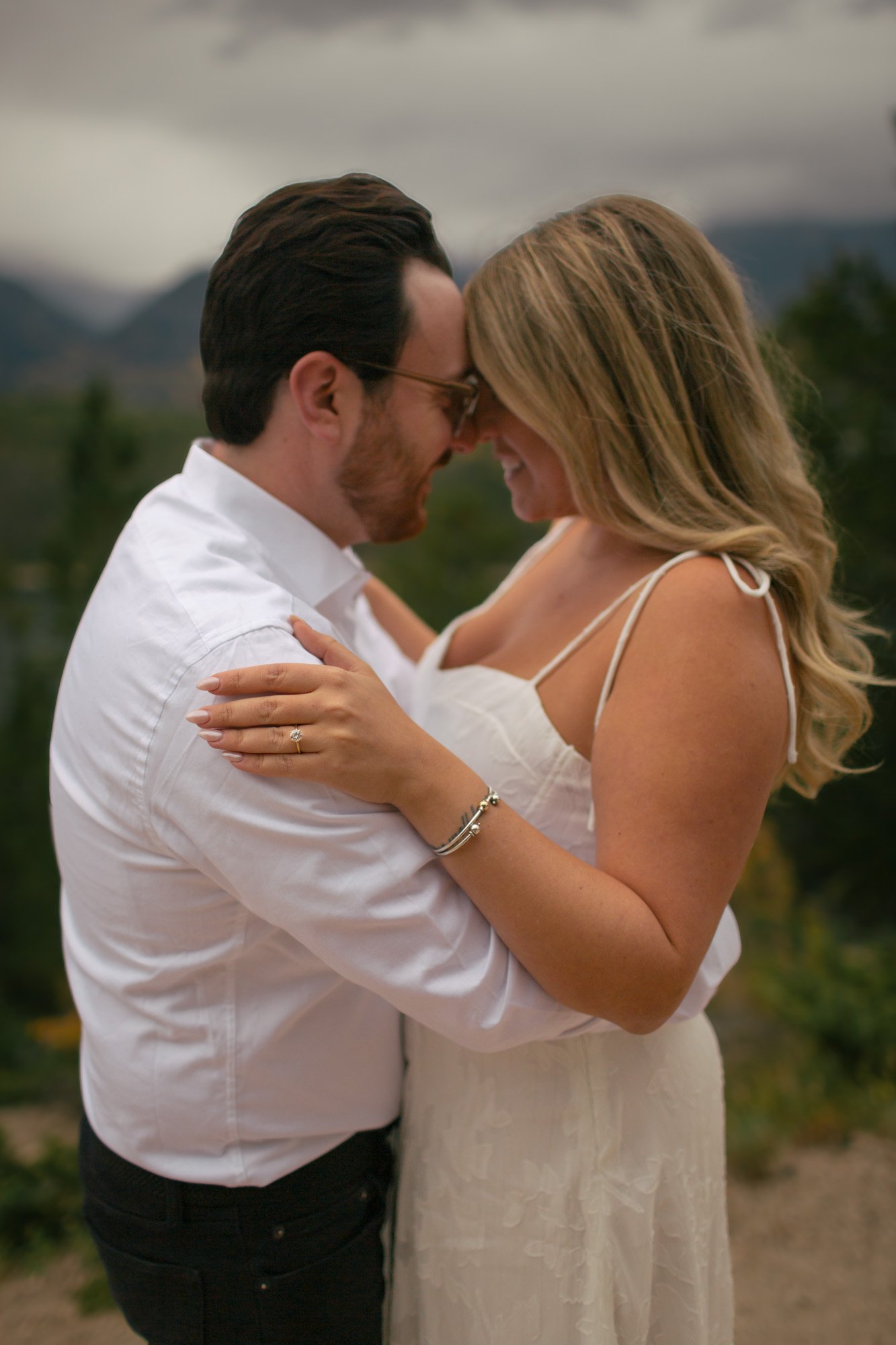 A man and woman are close together, touching foreheads and holding each other outdoors, with mountains and cloudy sky in the background.
