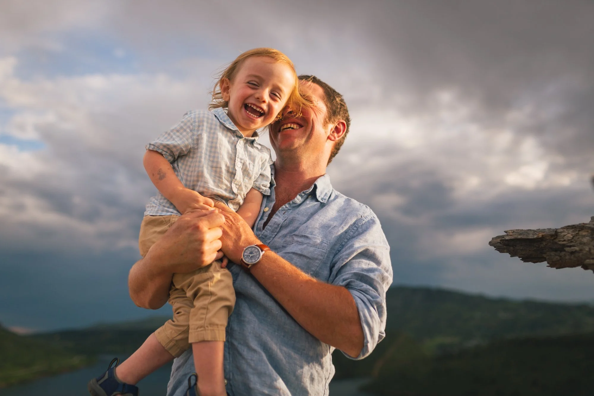 A man holding a young girl smiling with a cloudy sky and mountain landscape in the background.