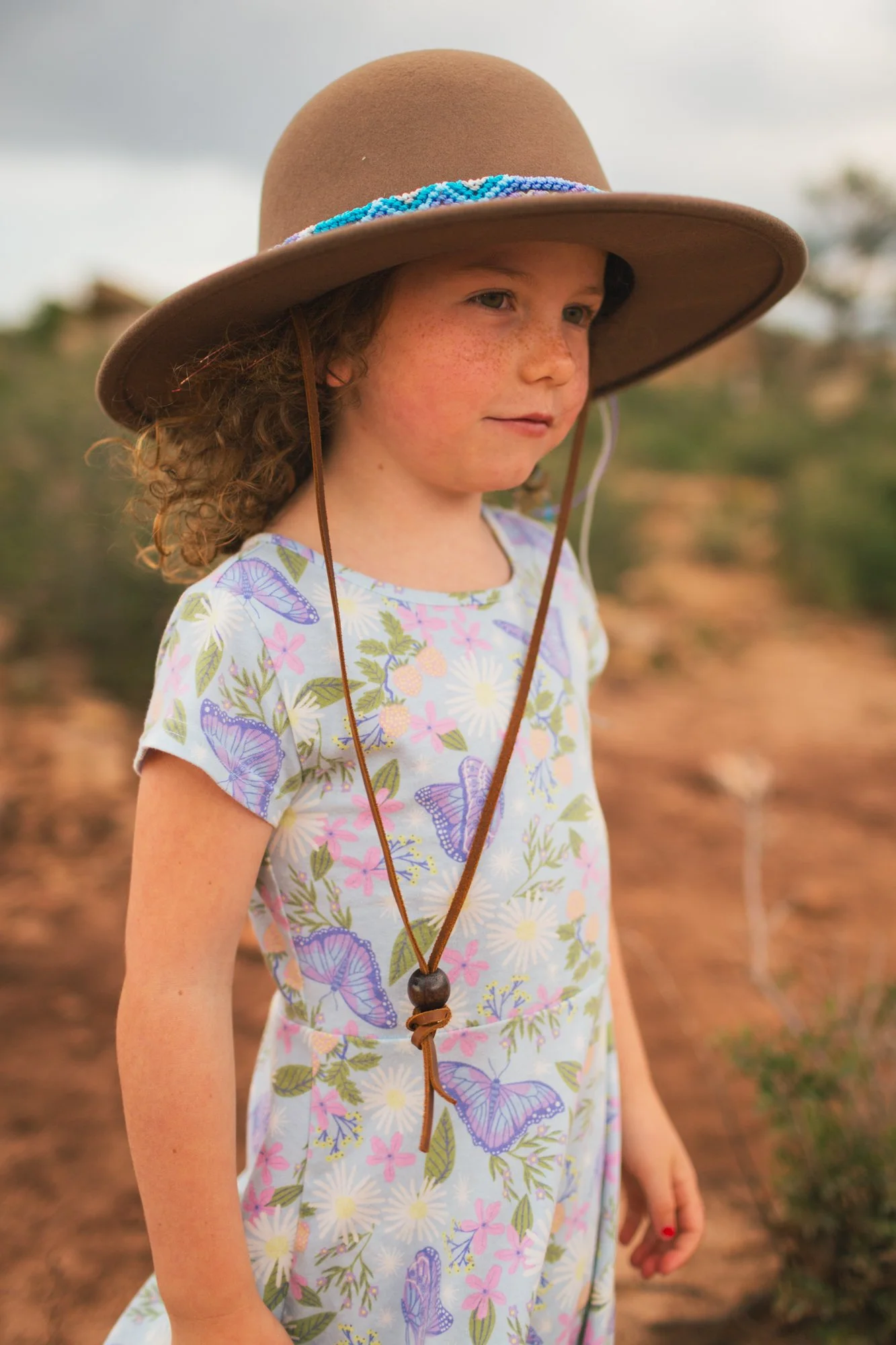 Young girl with curly hair and freckles wearing a beige wide-brimmed hat with blue beaded decoration, floral dress with butterflies, outdoors with blurred background.