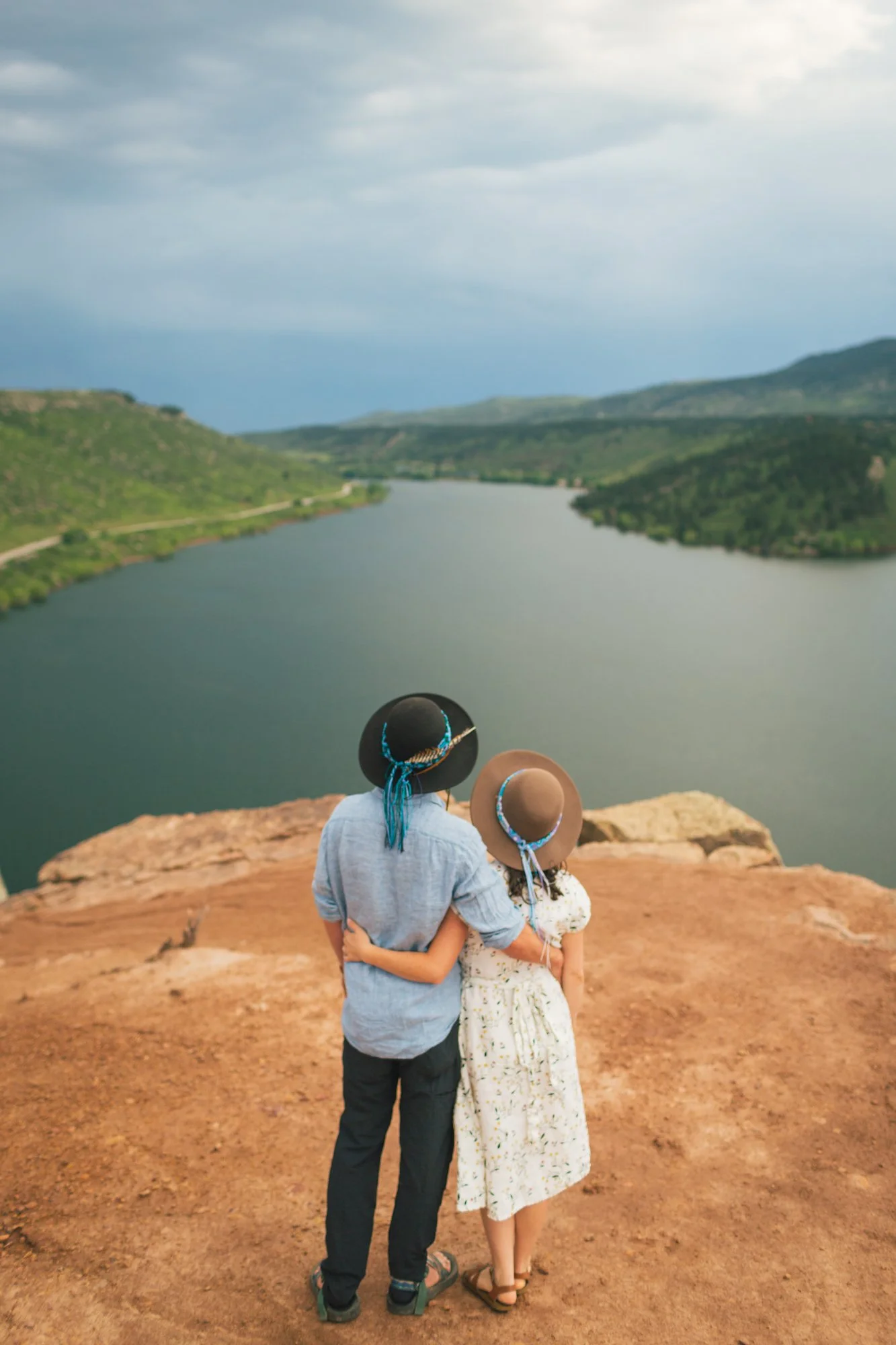 Two women dressed in white and blue with hats, standing closely together on a rocky ledge overlooking a river, with rolling hills and cloudy sky in the background.