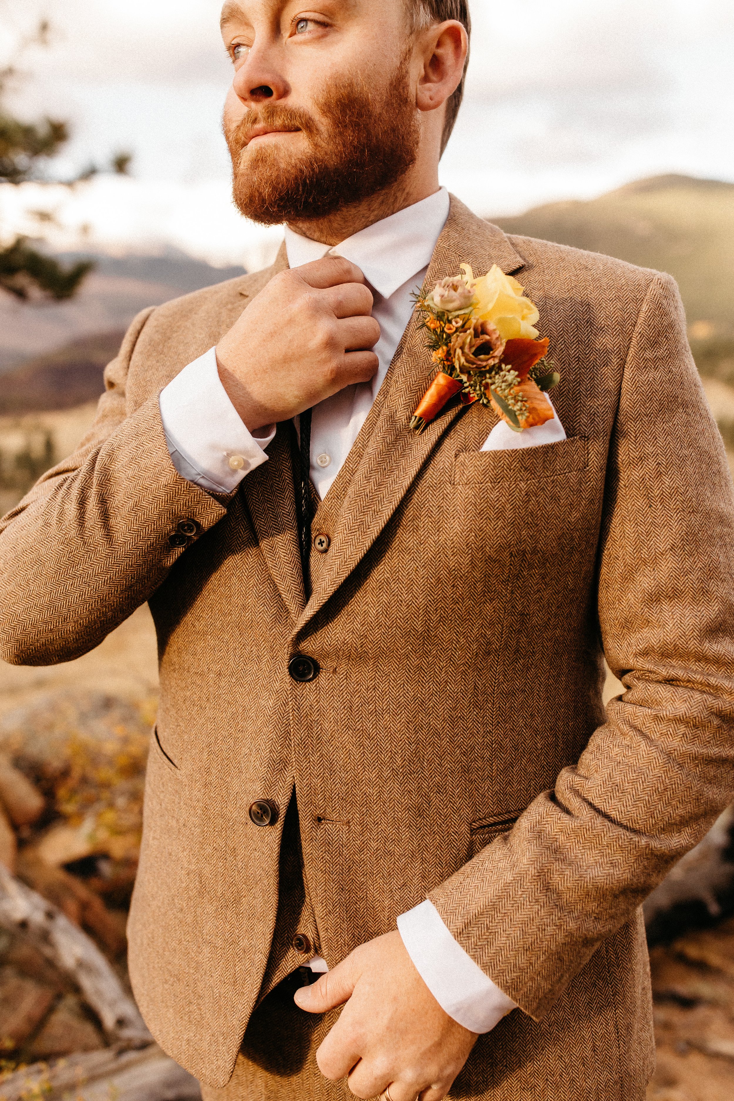 A man outdoors in a tan herringbone suit adjusting his white collared shirt with a boutonniere on his lapel, standing on a rocky landscape with mountains in the background.