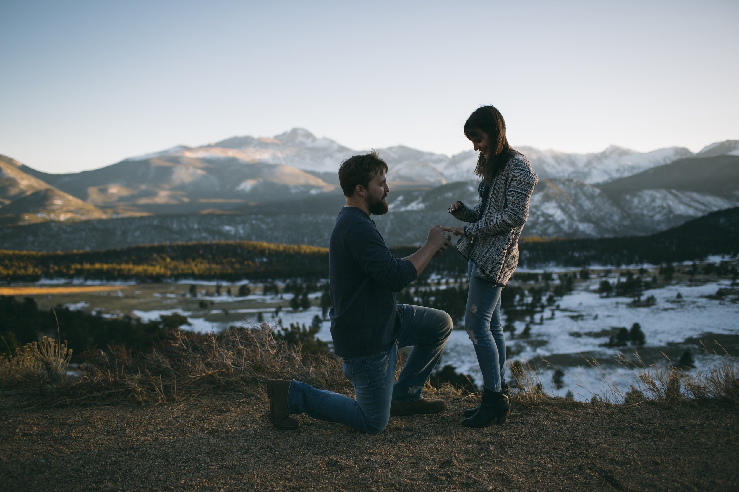 A man on one knee proposing to a woman outdoors against a mountain landscape and snow patches.