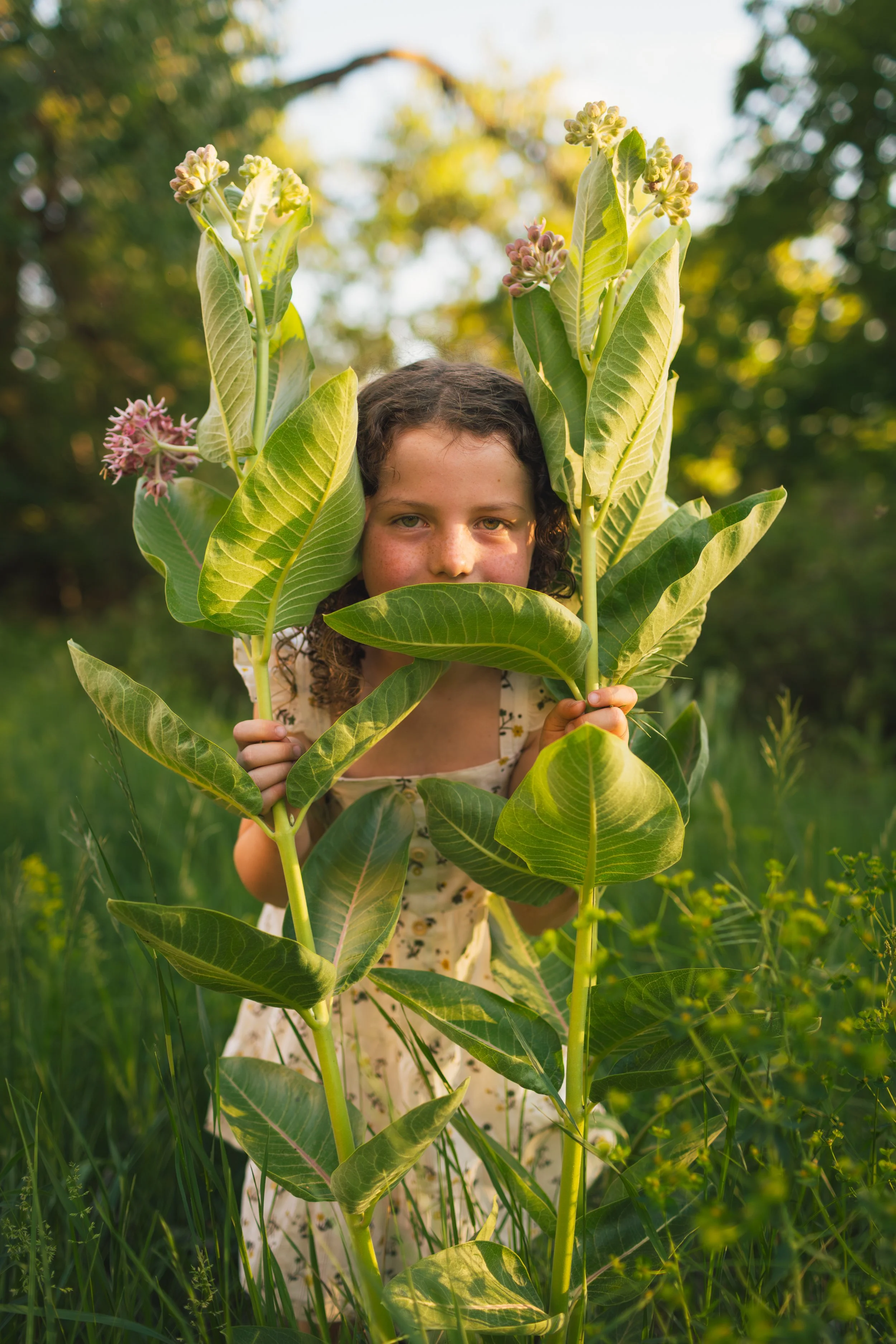 A young girl with curly hair and freckles, partially hidden behind large green leaves, holding a tall plant with pink flowers in a lush outdoor setting.
