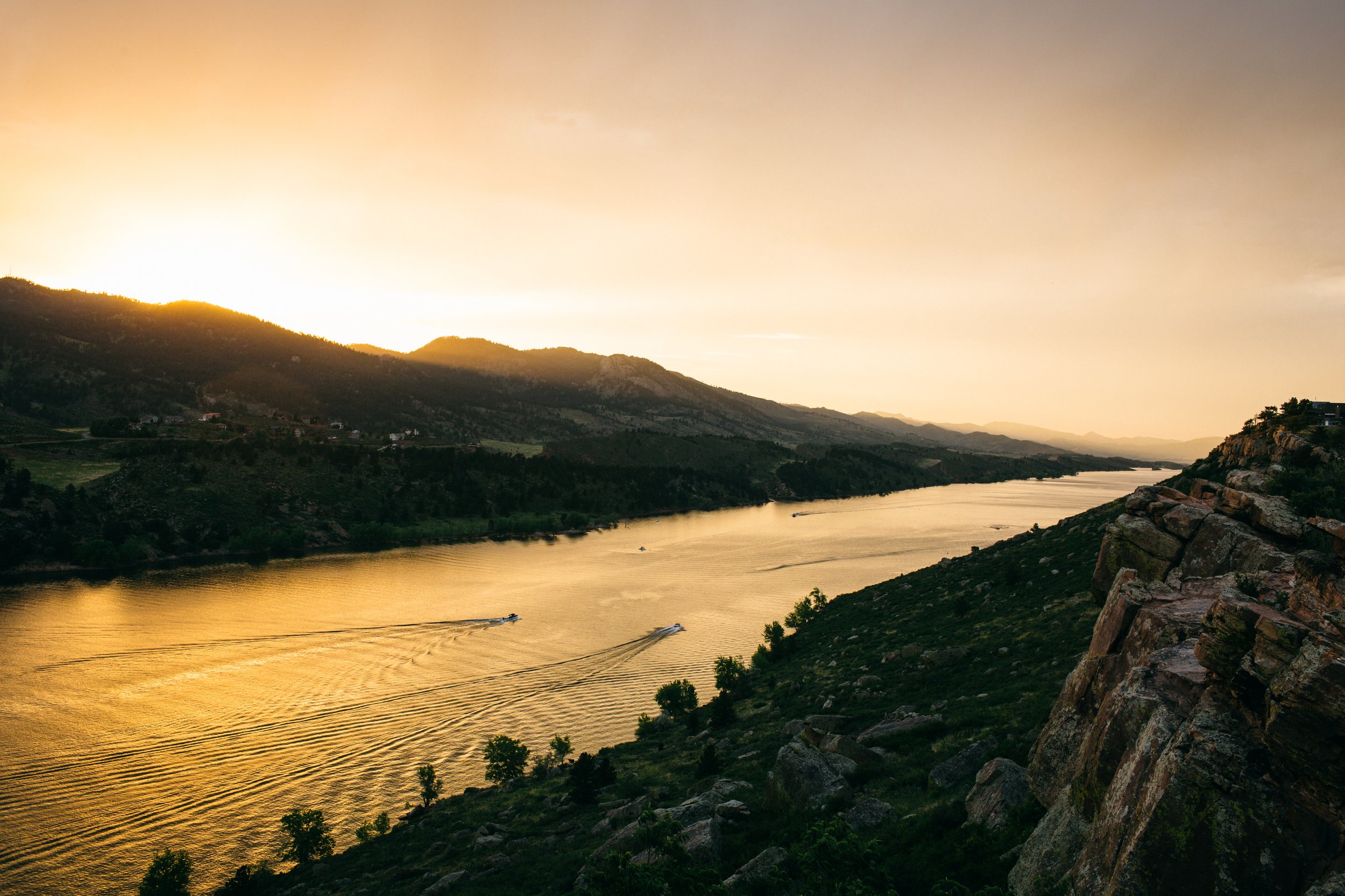 A river running through a mountainous landscape during sunset with boats on the water and rocks on the hillside.