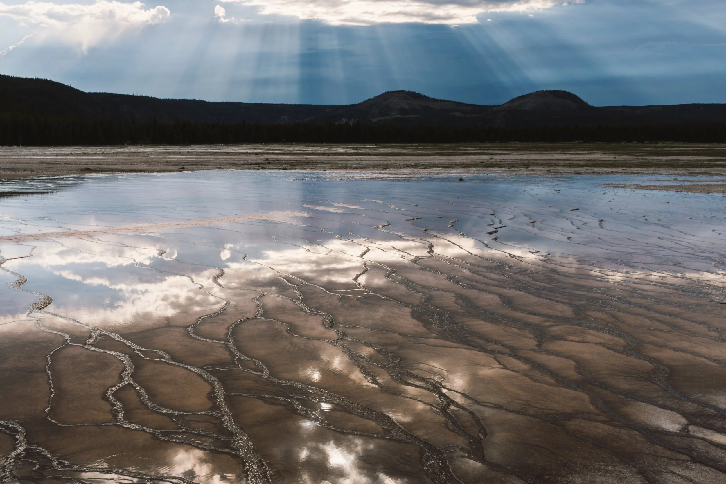 A landscape featuring a dry, cracked terrain with patches of water, a distant forest, mountain hills in the background, and a partly cloudy sky with rays of sunlight shining through.