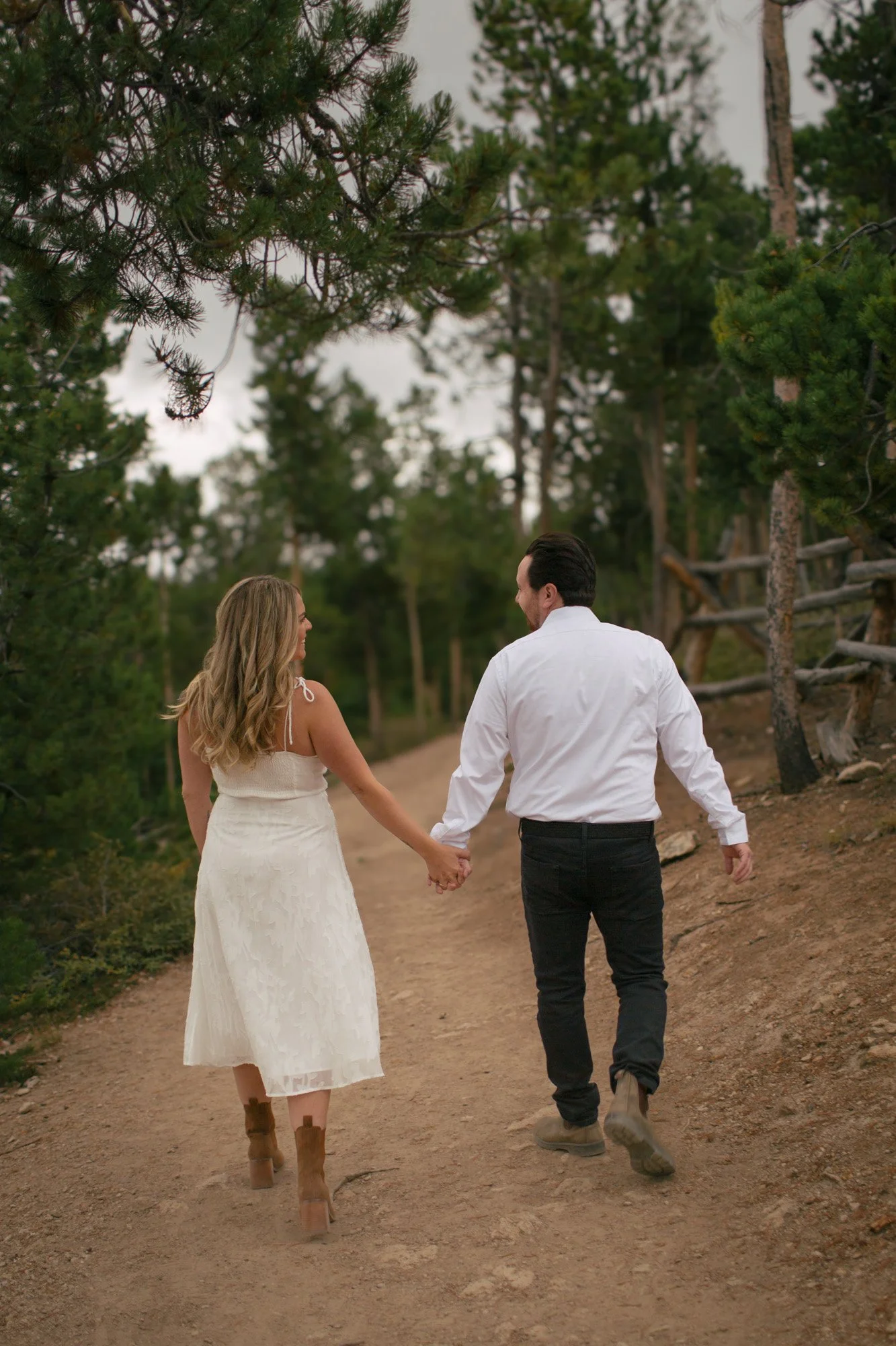 A couple, a woman in a white dress and boots and a man in a white shirt and dark pants, walking hand in hand on a dirt path surrounded by trees.
