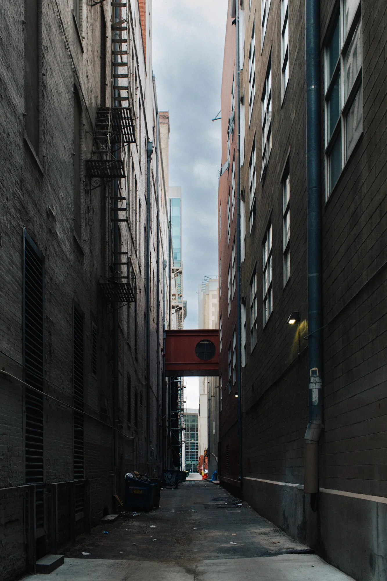 An alley between two tall buildings with fire escapes on the left side and drainage pipes on the right, leading to a street in the distance under a cloudy sky.