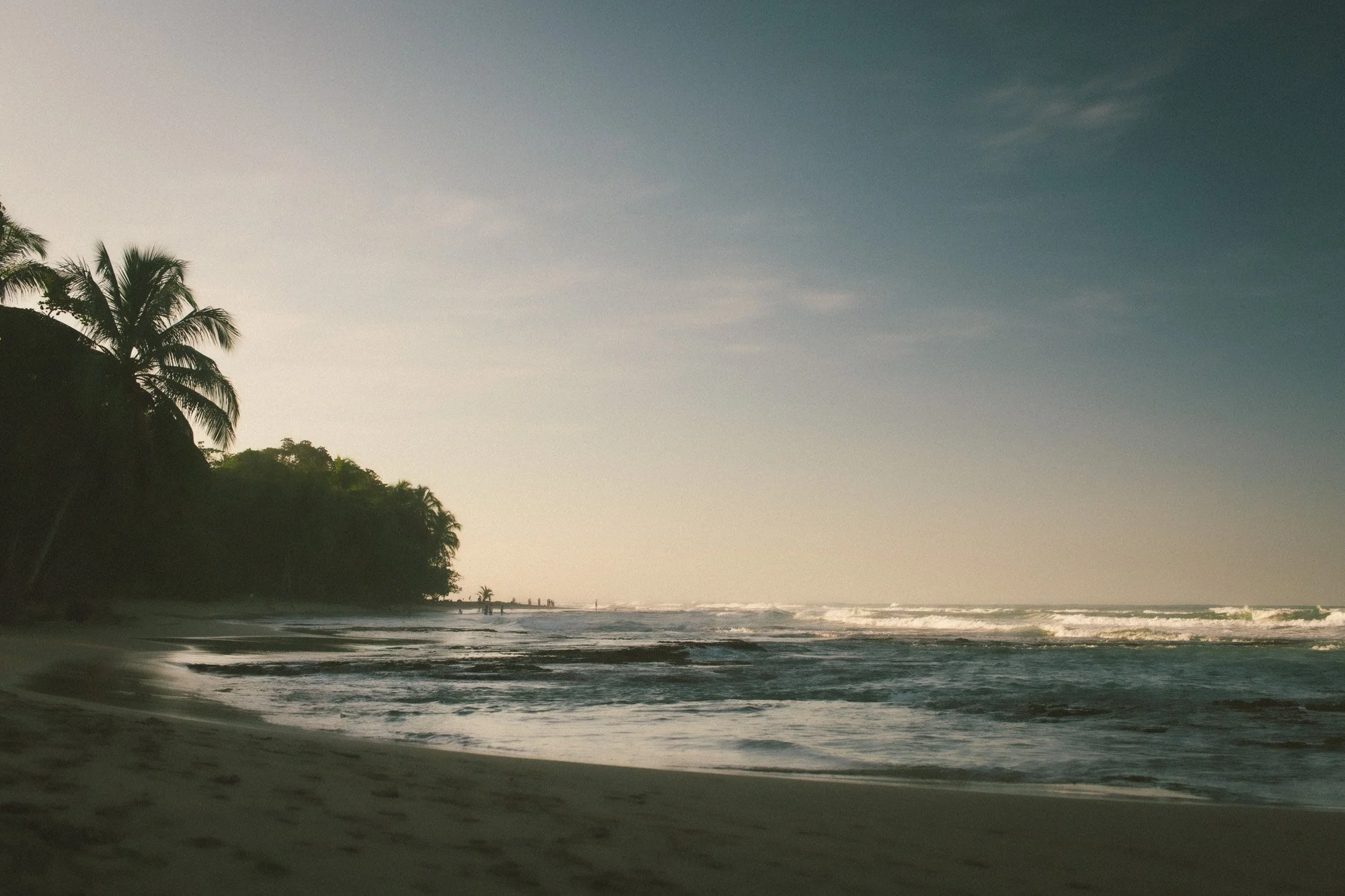 A sunset or sunrise at a tropical beach with palm trees, calm ocean waves, and a few people walking along the shoreline.