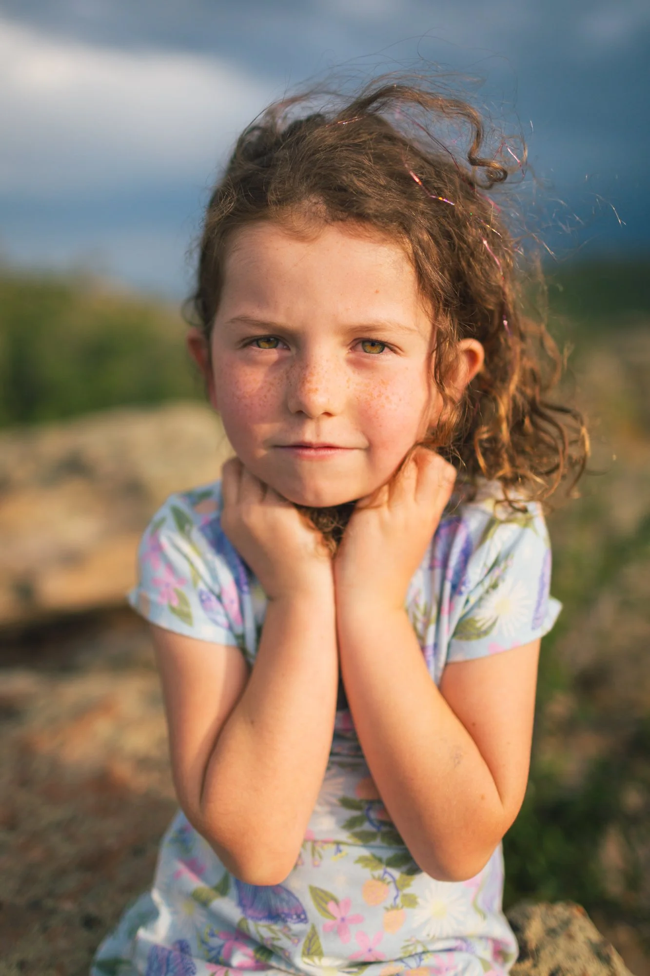 A young girl with curly brown hair and freckles, wearing a floral dress, holding her hands near her face, outdoors during late afternoon or early evening with a cloudy sky in the background.