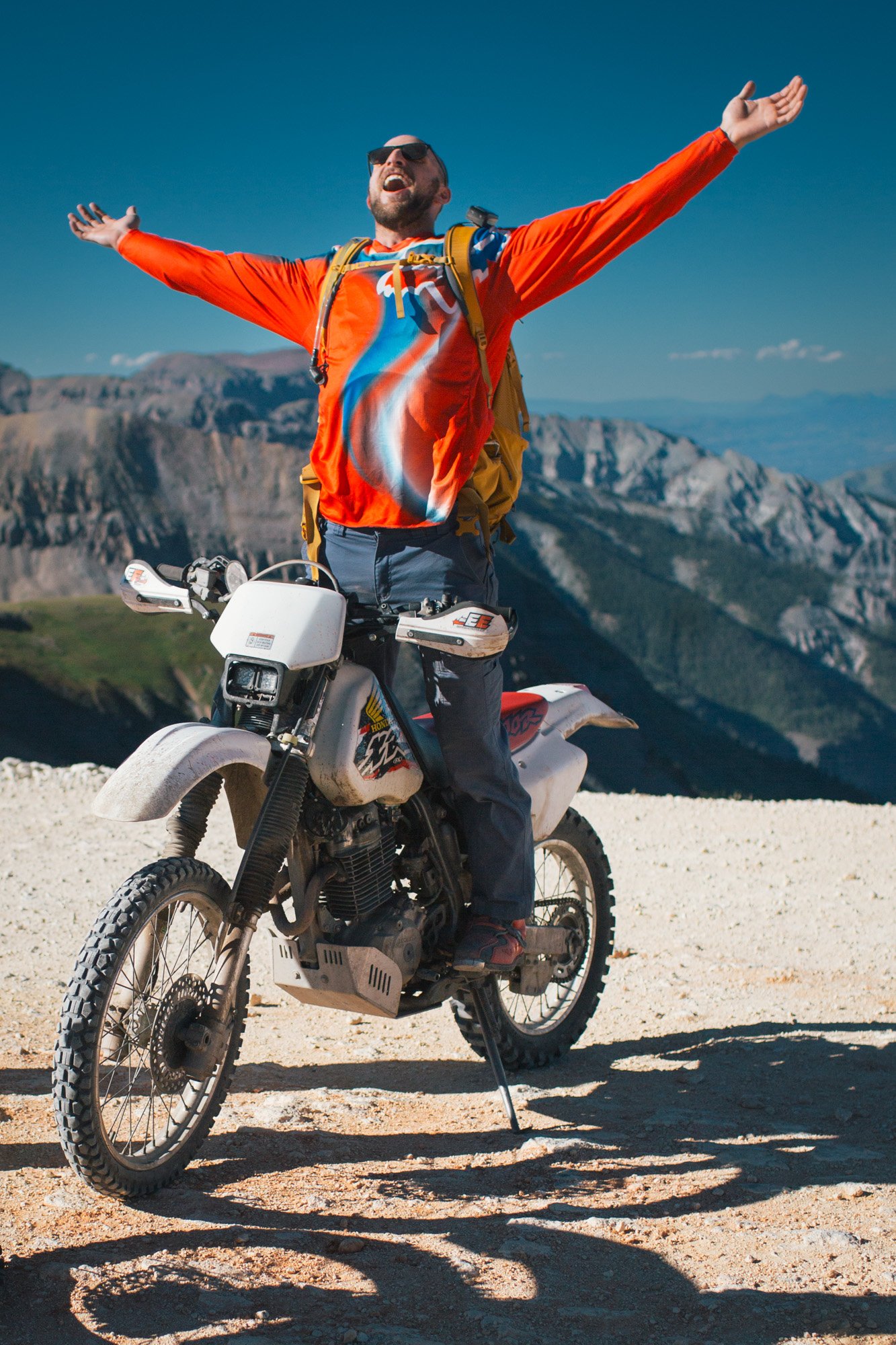 A man wearing sunglasses, a bright orange jacket, and a colorful shirt stands on a dirt path with his arms outstretched, smiling with his head tilted back. He is standing on a dirt bike with a mountainous landscape in the background under a clear blu