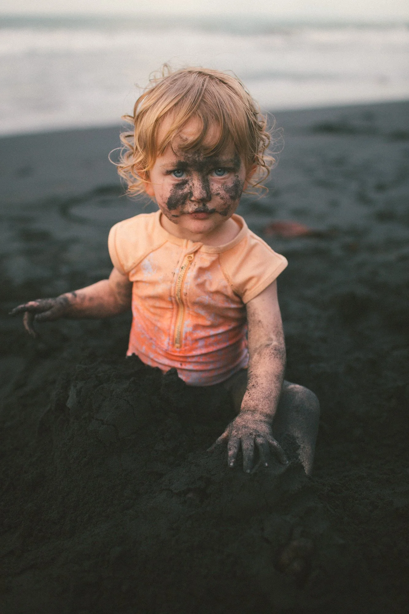 A young child with blonde curly hair and blue eyes, covered in mud, sitting on black sand at the beach, with water and waves in the background.