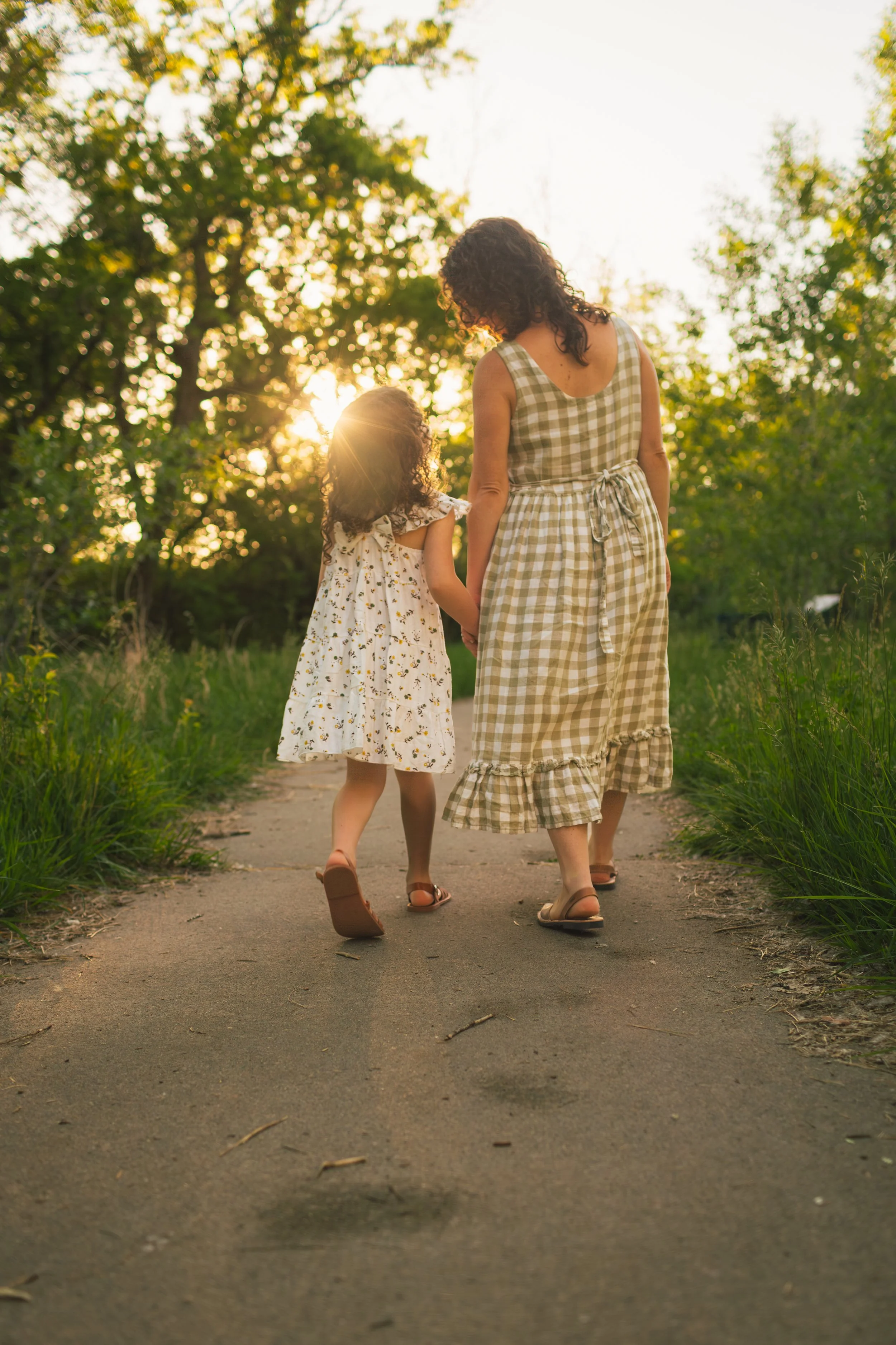 A woman and a young girl are walking hand in hand along a sidewalk surrounded by greenery. The woman is wearing a checkered dress with a belt, and the girl is in a floral dress. The sun is setting behind trees, creating a warm glow.