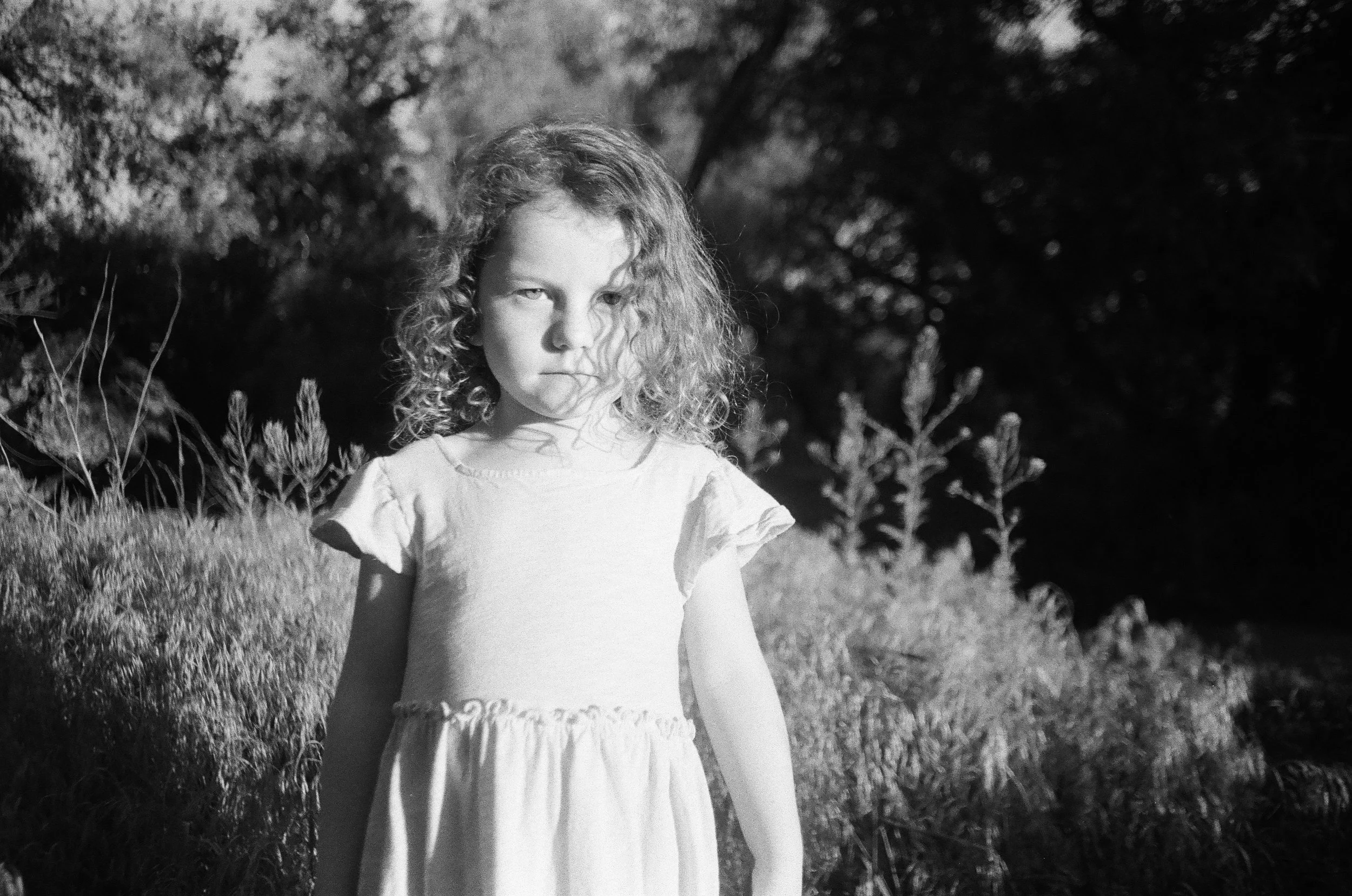 A young girl with curly hair standing outdoors in a grassy area near trees, wearing a light-colored dress, in black and white.