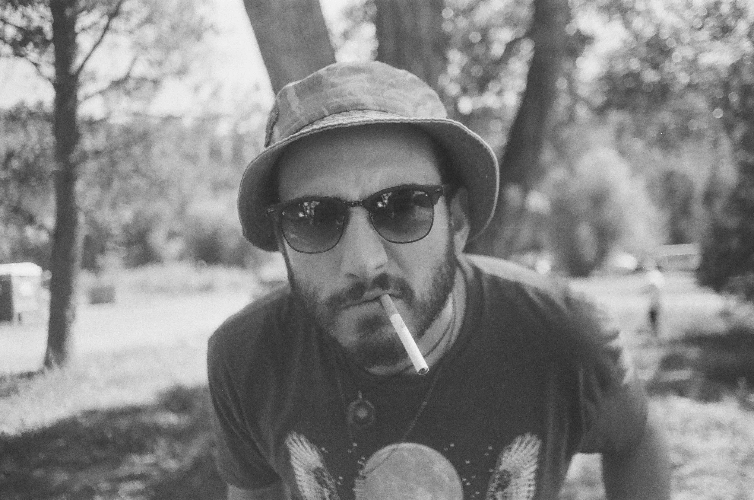 A person wearing a patterned beanie, dark sunglasses, and a jacket, smoking a cigarette outdoors with a blurred background of trees.