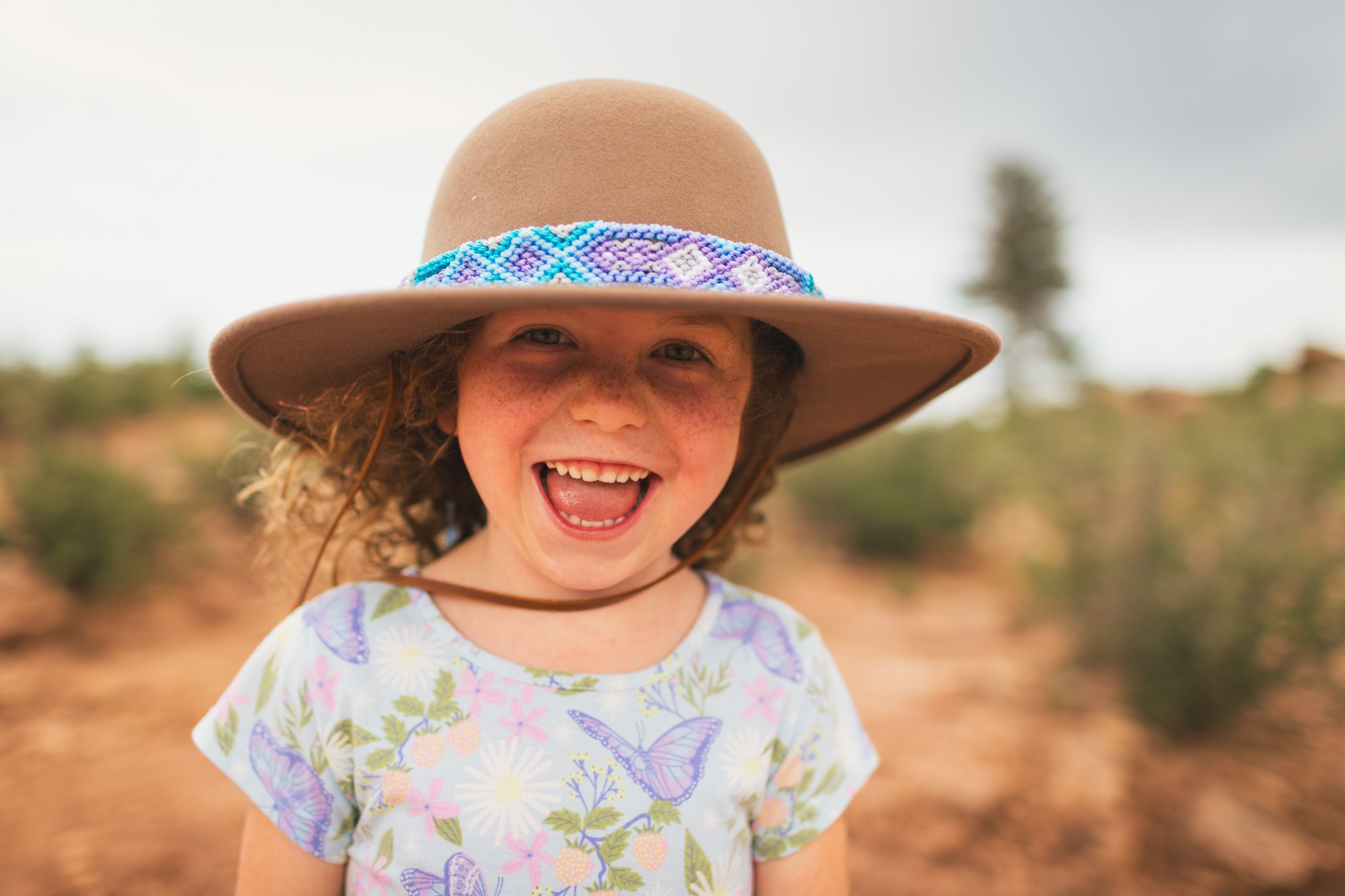 A young girl with curly hair smiling outdoors, wearing a large tan hat with a colorful beaded band and a shirt with butterfly and flower patterns.