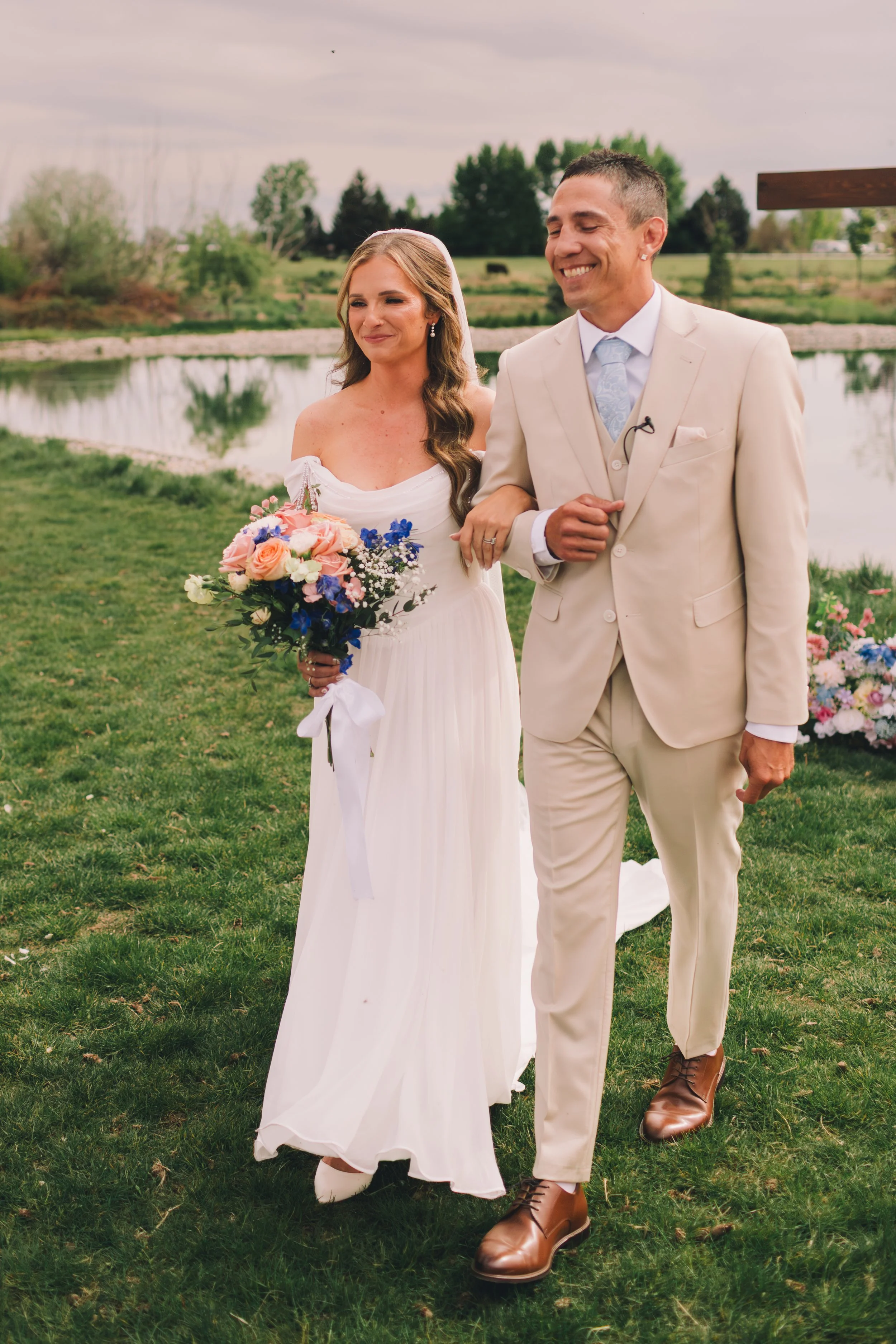 A newlywed couple walking arm-in-arm outdoors by a pond, with the woman holding a bouquet of pink, white, and purple flowers. The bride is wearing a white wedding dress, and the groom is dressed in a beige suit with brown shoes. They are smiling and 