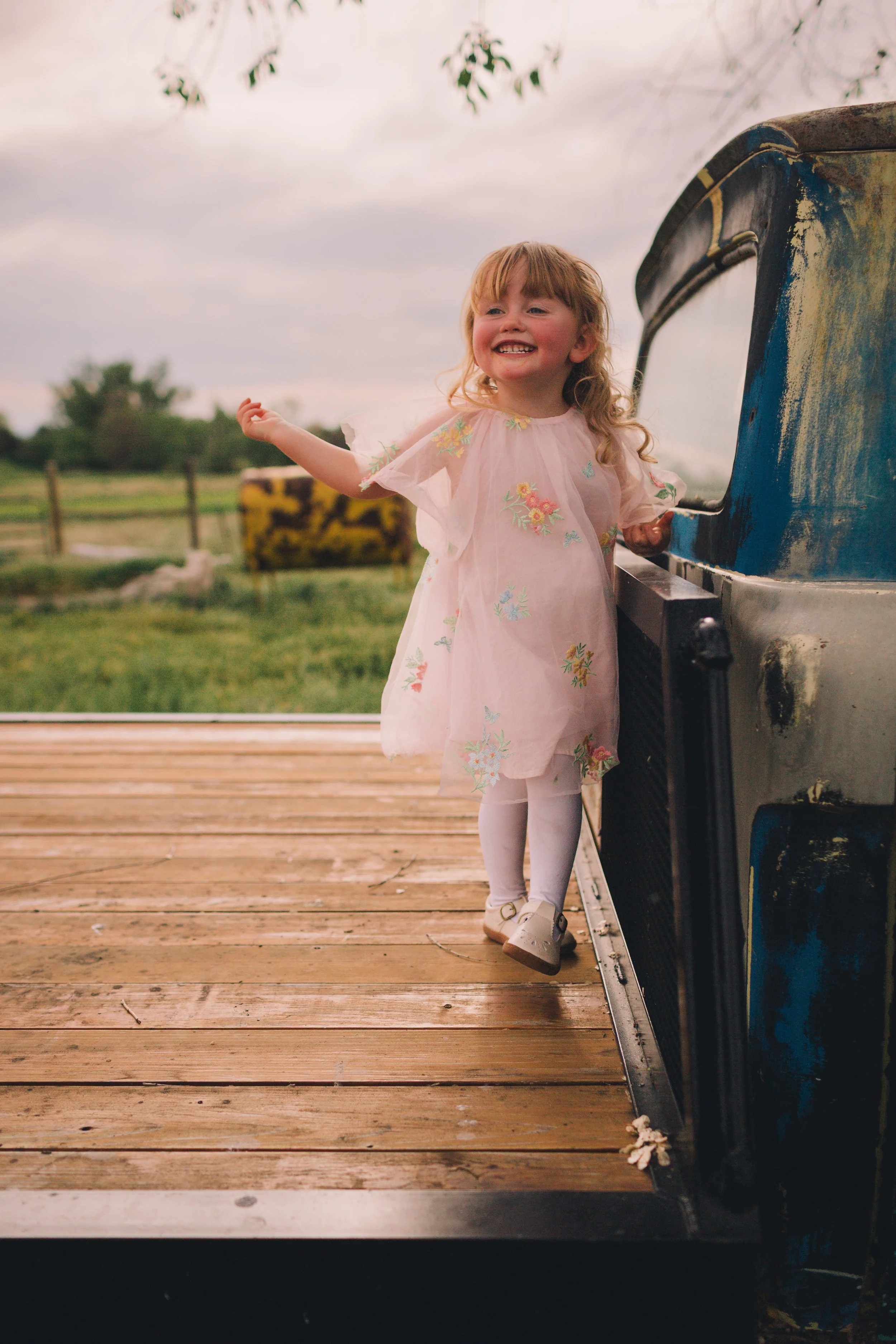 A young girl with blonde curly hair smiling and wearing a light pink dress with floral embroidery, standing next to an ice cream truck on a wooden platform outdoors during daytime with a cloudy sky.