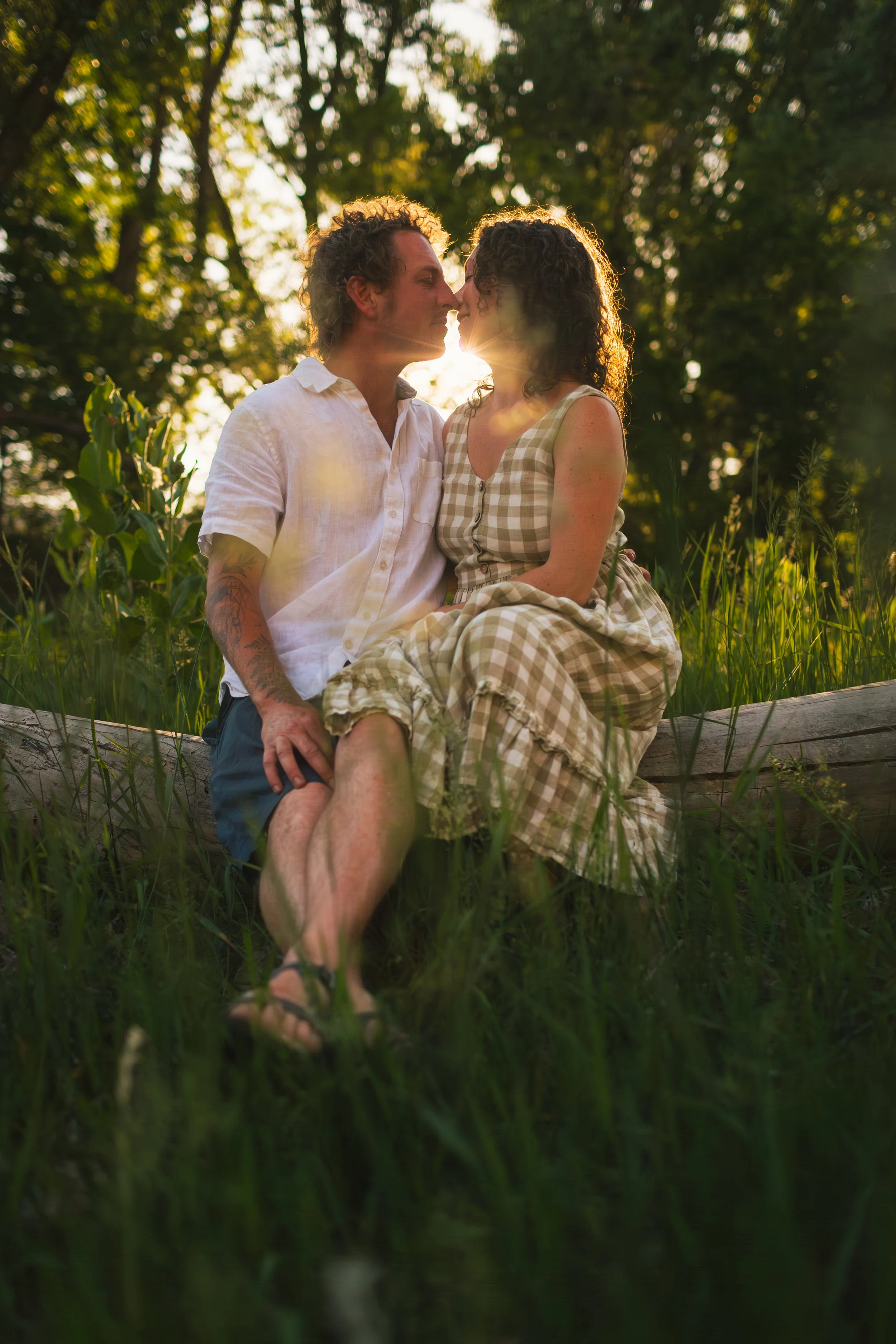 A couple sitting on a wooden log outdoors during sunset, nose to nose, in a green grassy area with trees in the background.