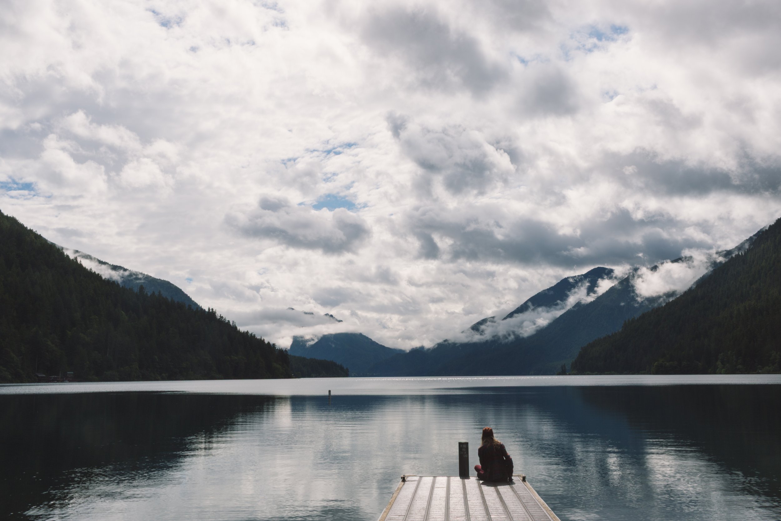 A person sitting on a dock by a calm lake, surrounded by mountains with clouds overhead.