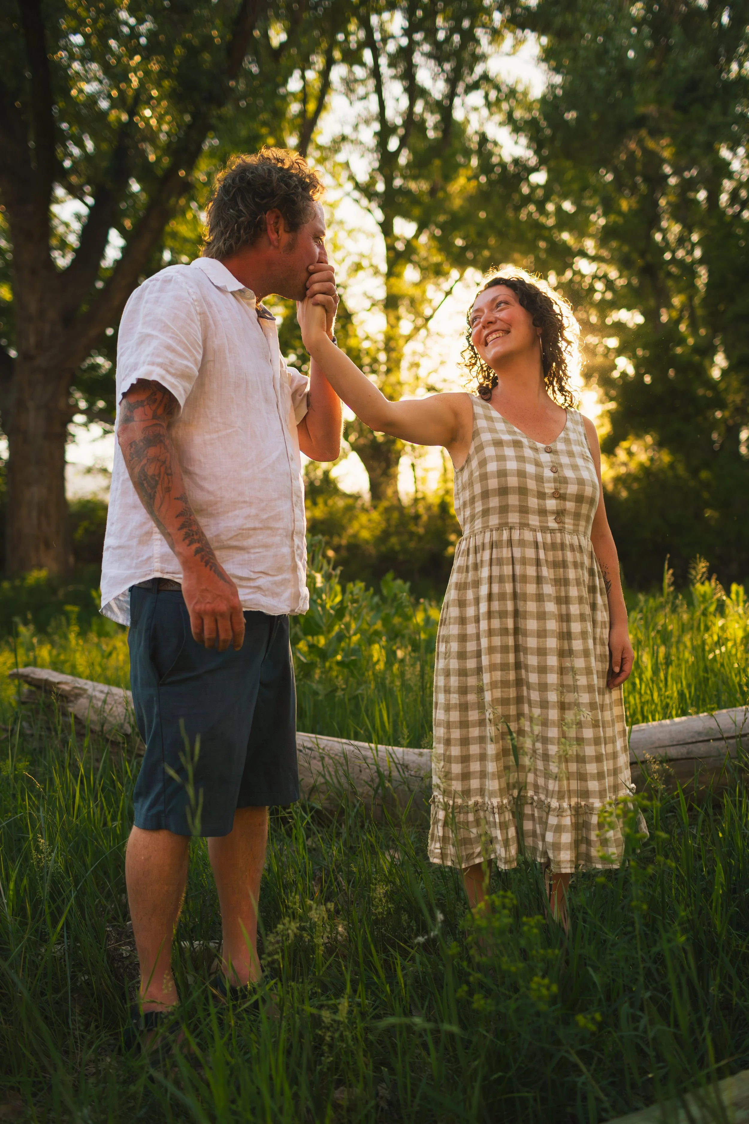 A man and woman standing outdoors in a grassy field with tall trees behind them. The woman is touching the man's face, and they are smiling at each other. The sunlight filters through the leaves, creating a warm, golden glow.