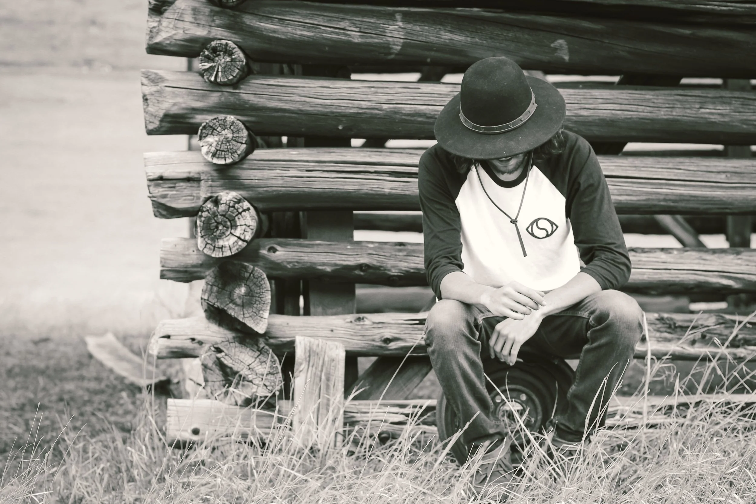 A person wearing a wide-brimmed hat and a black and white long-sleeve shirt with a logo, sitting on grass in front of a stacked log wall, looking down with arms crossed and hands clasped, in a black and white photograph.