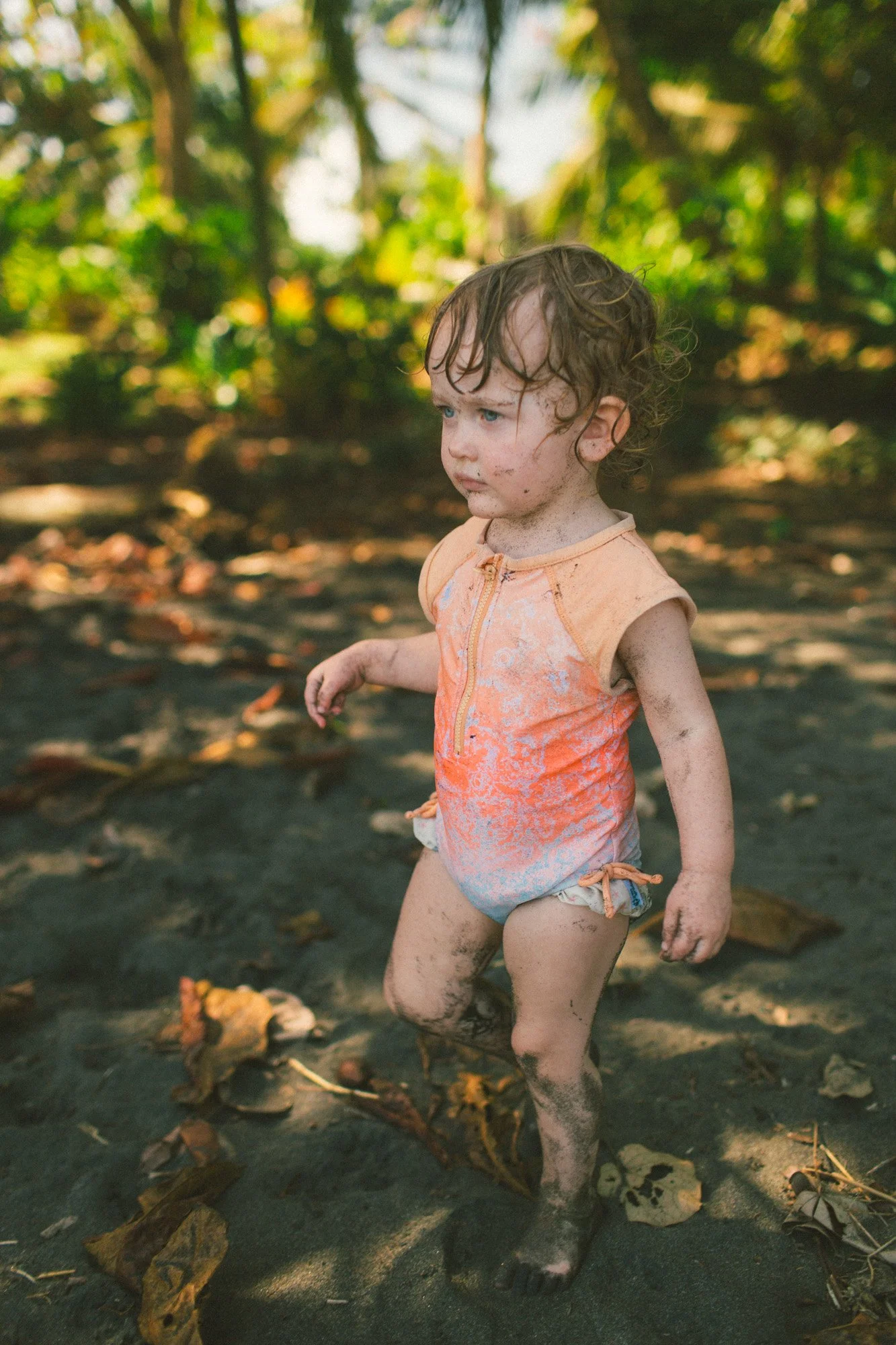 Young child with dirty face and body, wearing a sleeveless orange shirt and diaper, standing outdoors on a dirt surface with fallen leaves and trees in the background.