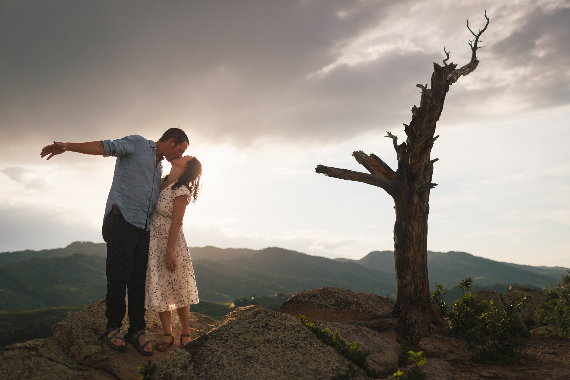 A couple kissing on a rocky hilltop near a leafless tree with mountains in the background during sunset.