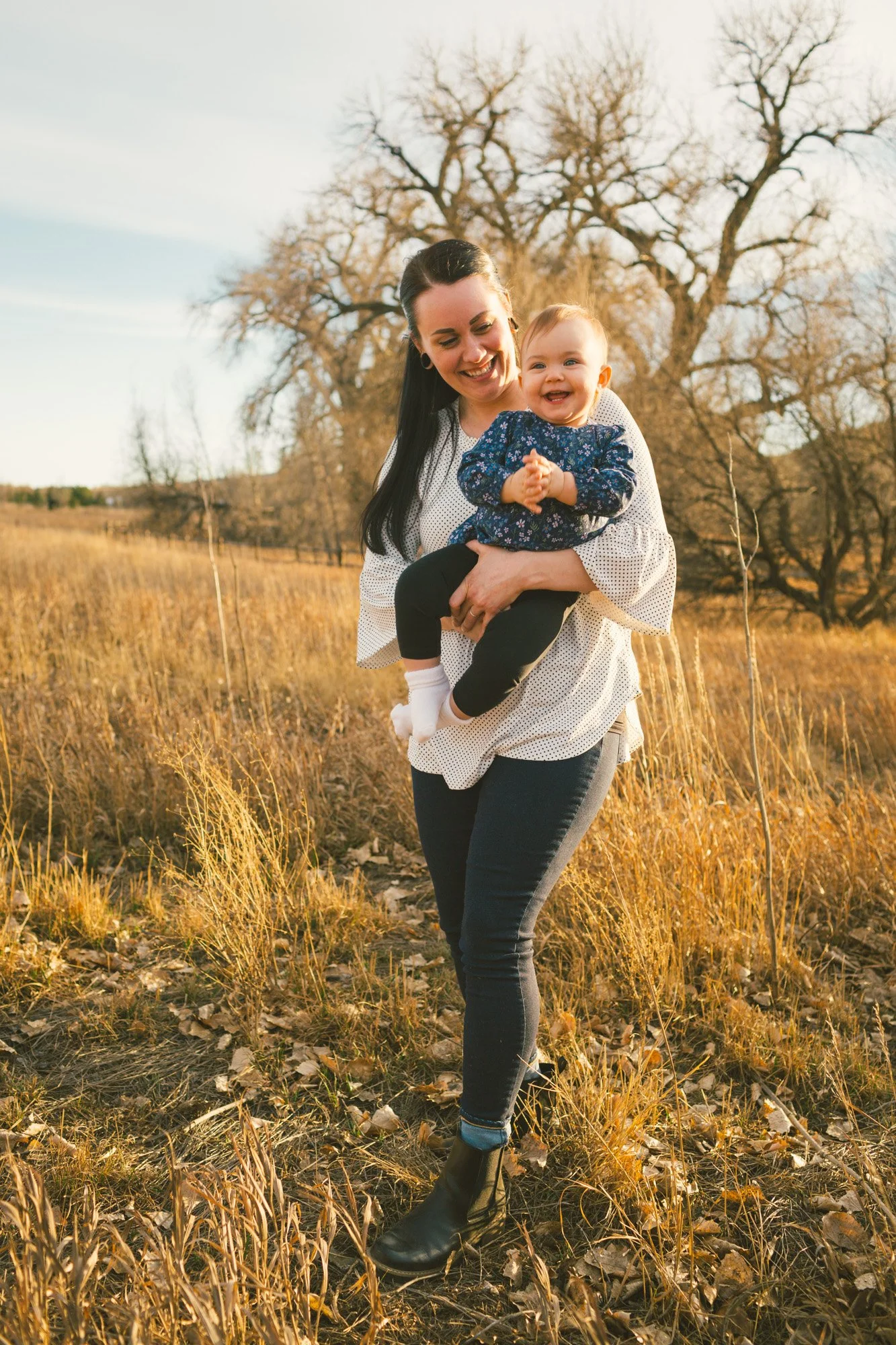 A woman with dark hair smiling and holding a cheerful baby girl with blonde hair in an outdoor field with golden grass and leafless trees in the background during autumn.