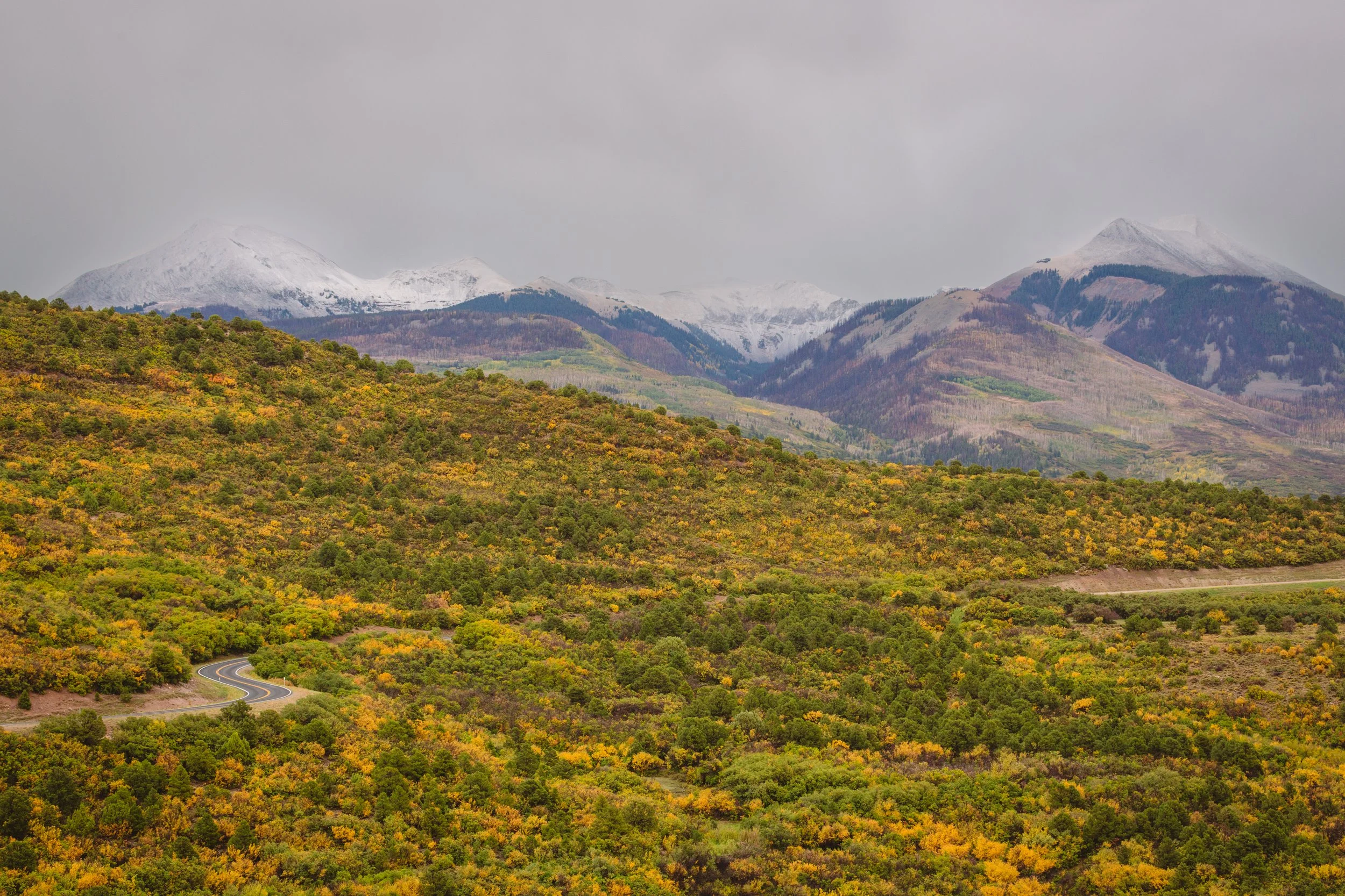 Mountain range with snow-capped peaks and a winding road through a forested landscape with fall foliage.