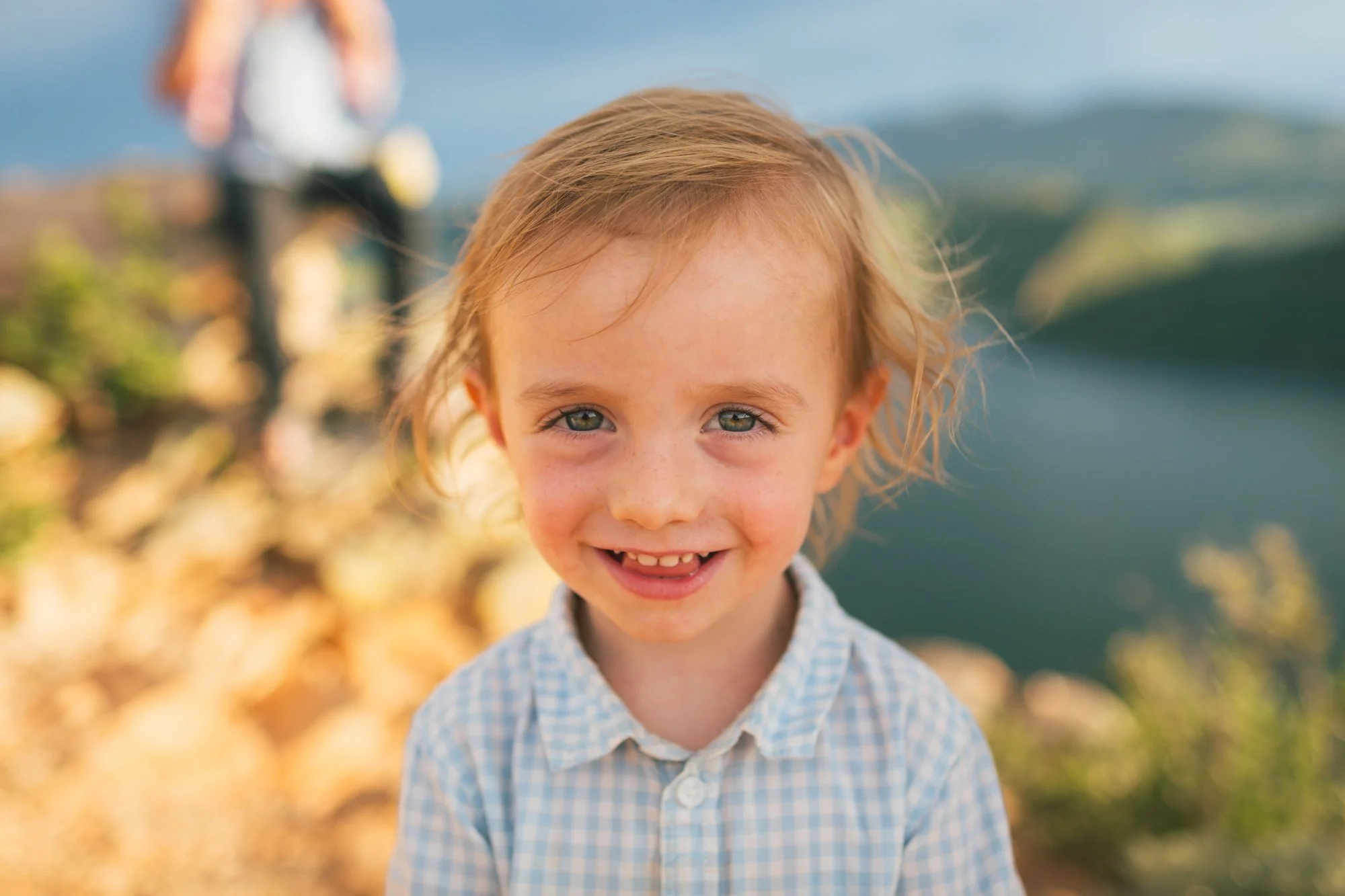 A young boy with light brown, curly hair and blue eyes smiling outdoors with blurred autumn leaves and landscape in the background.