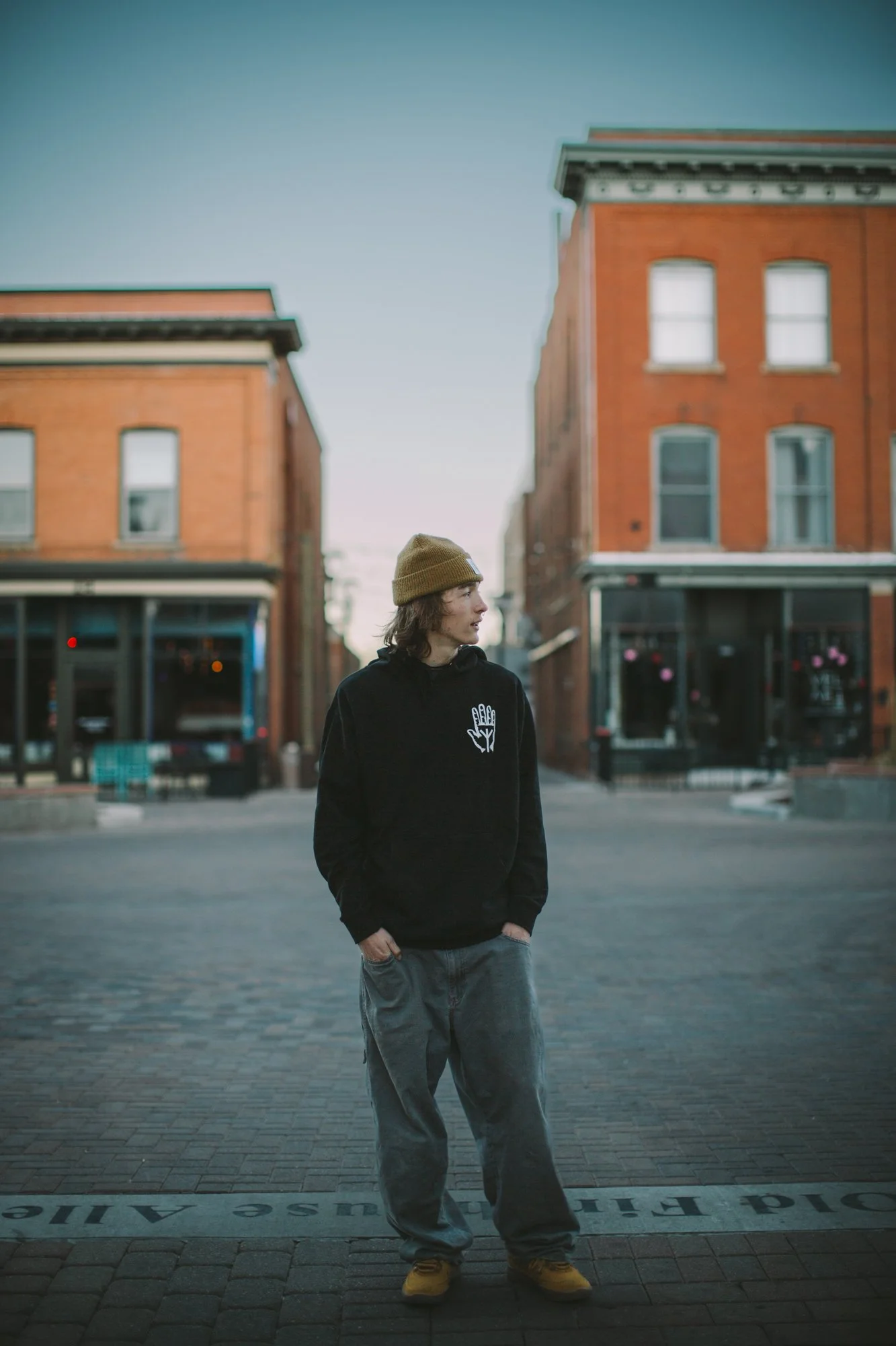 A young man with long hair, wearing a yellow knit cap, black hoodie with a white hand symbol, gray sweatpants, and yellow shoes, standing on a paved street with brick buildings in the background and a clear sky overhead.
