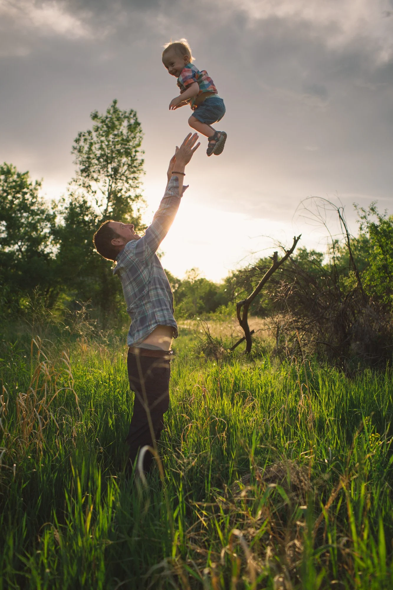 Man lifting a young boy into the air outdoors during sunset in a grassy field with trees.