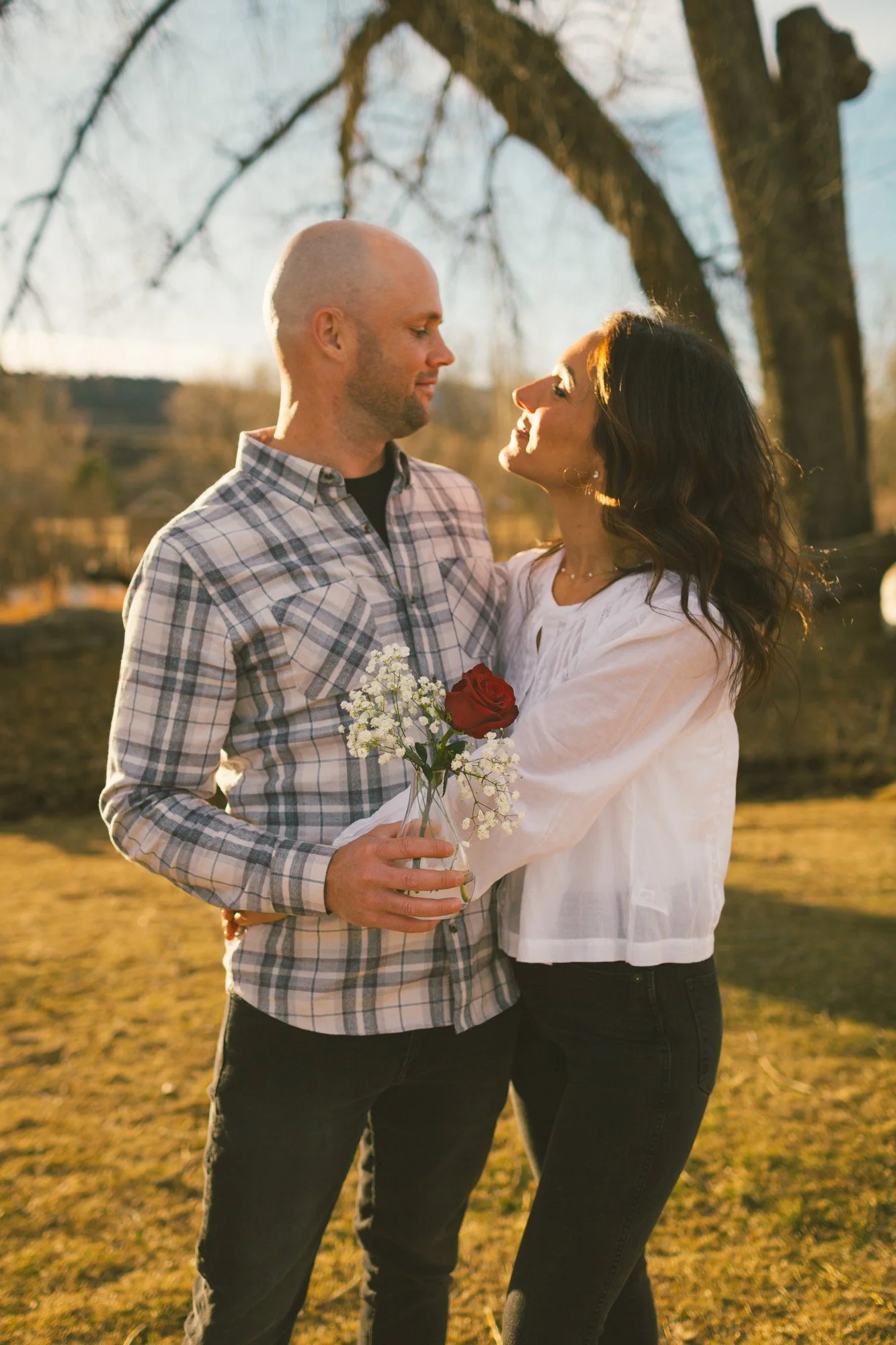 A couple outdoors in the late afternoon, holding a bouquet of flowers, with the man looking at the woman in front of a tree and a clear sky.