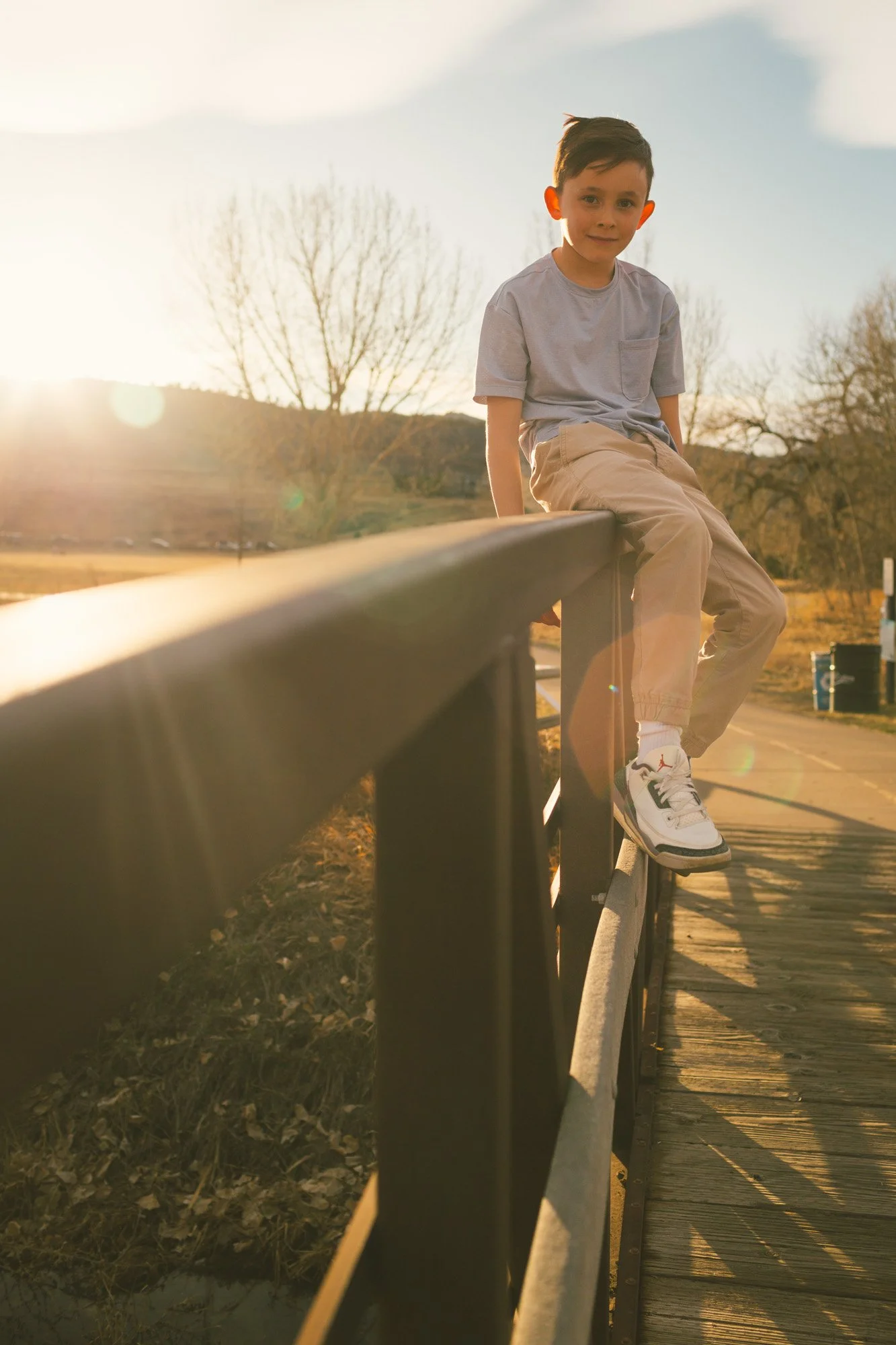 A young boy sitting on a wooden railing during sunset, wearing a light gray t-shirt, beige pants, and white sneakers, with leafless trees and a hilly landscape in the background.
