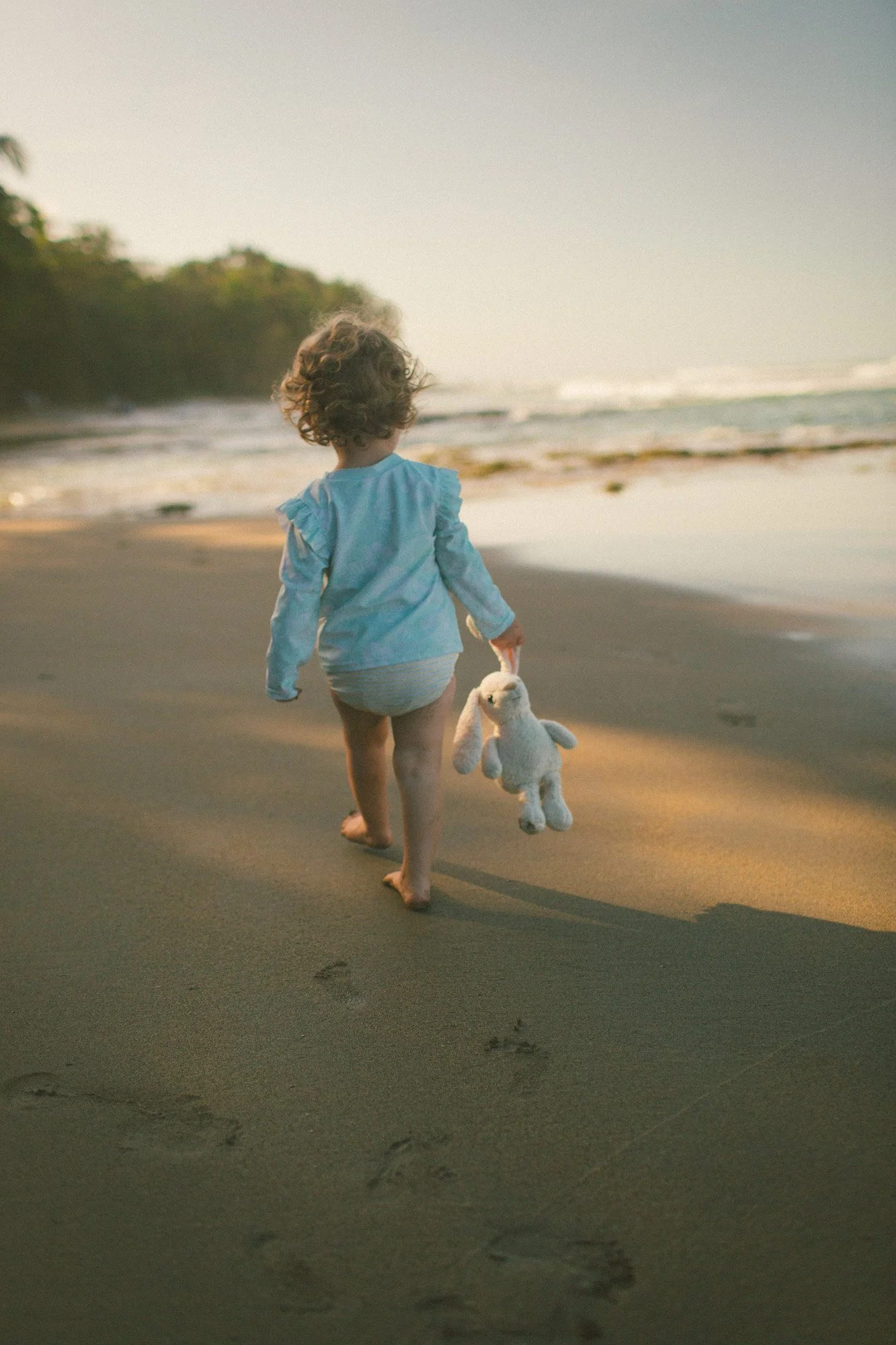 A young girl walking on the beach at sunset, holding a stuffed bunny toy, with the ocean and a distant tree line in the background.