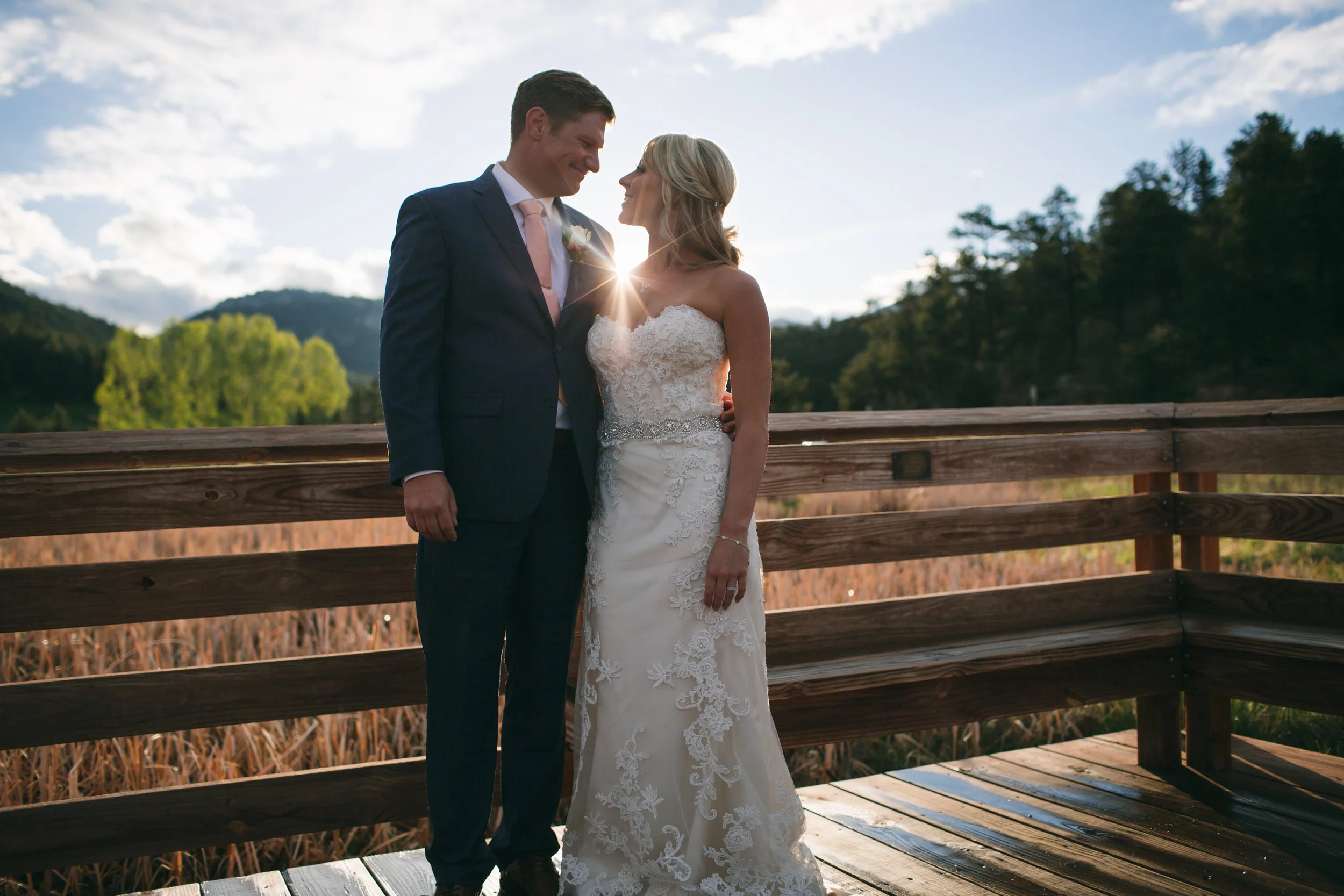 A newlywed couple stands close together on a wooden bridge outdoors, with the setting sun behind them. They are smiling and gazing into each other's eyes, surrounded by a scenic landscape of trees, mountains, and a partly cloudy sky.