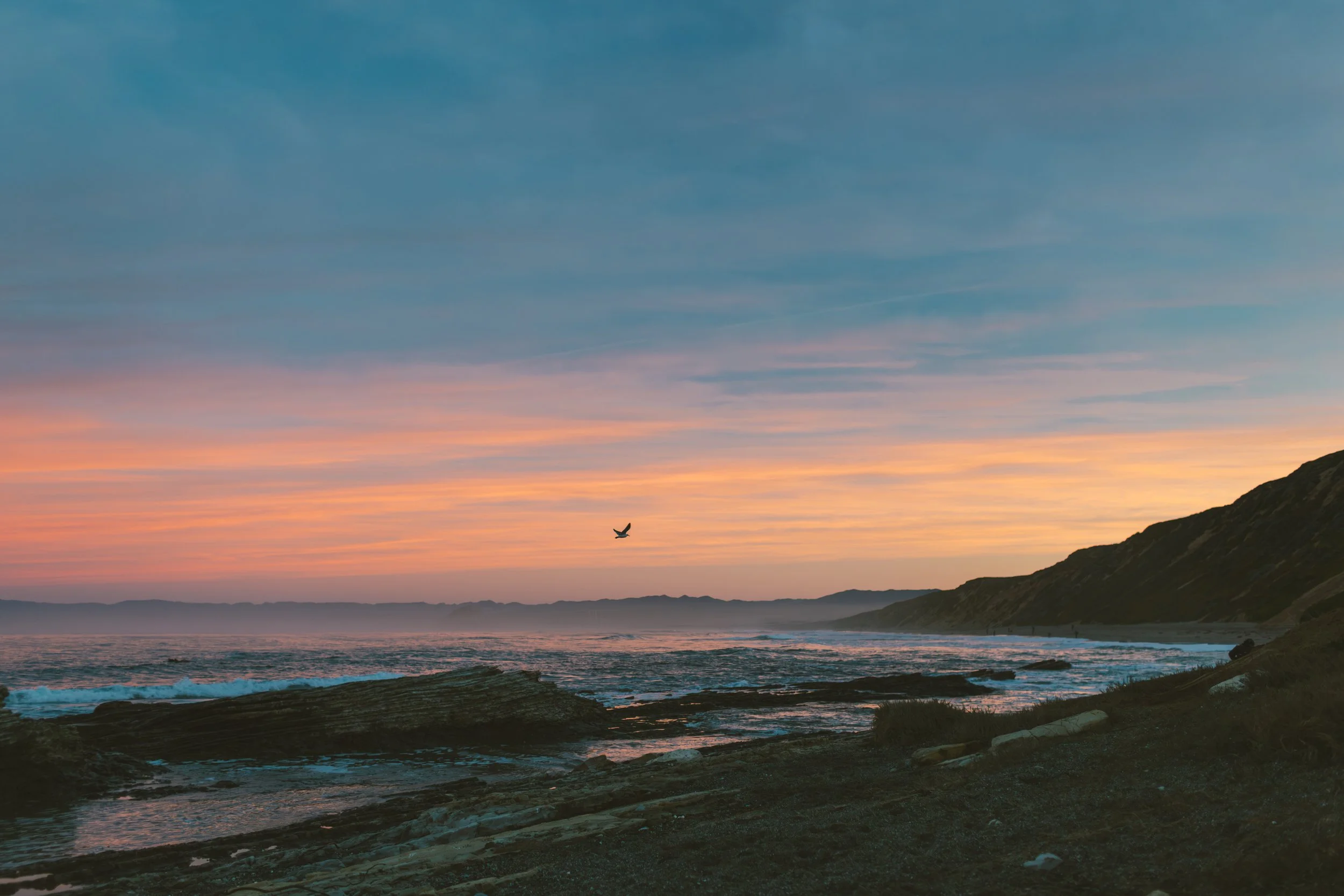 Scenic view of the ocean at sunset with a bird flying in the sky, rocky shoreline in the foreground, and mountains in the distance under a colorful sky.