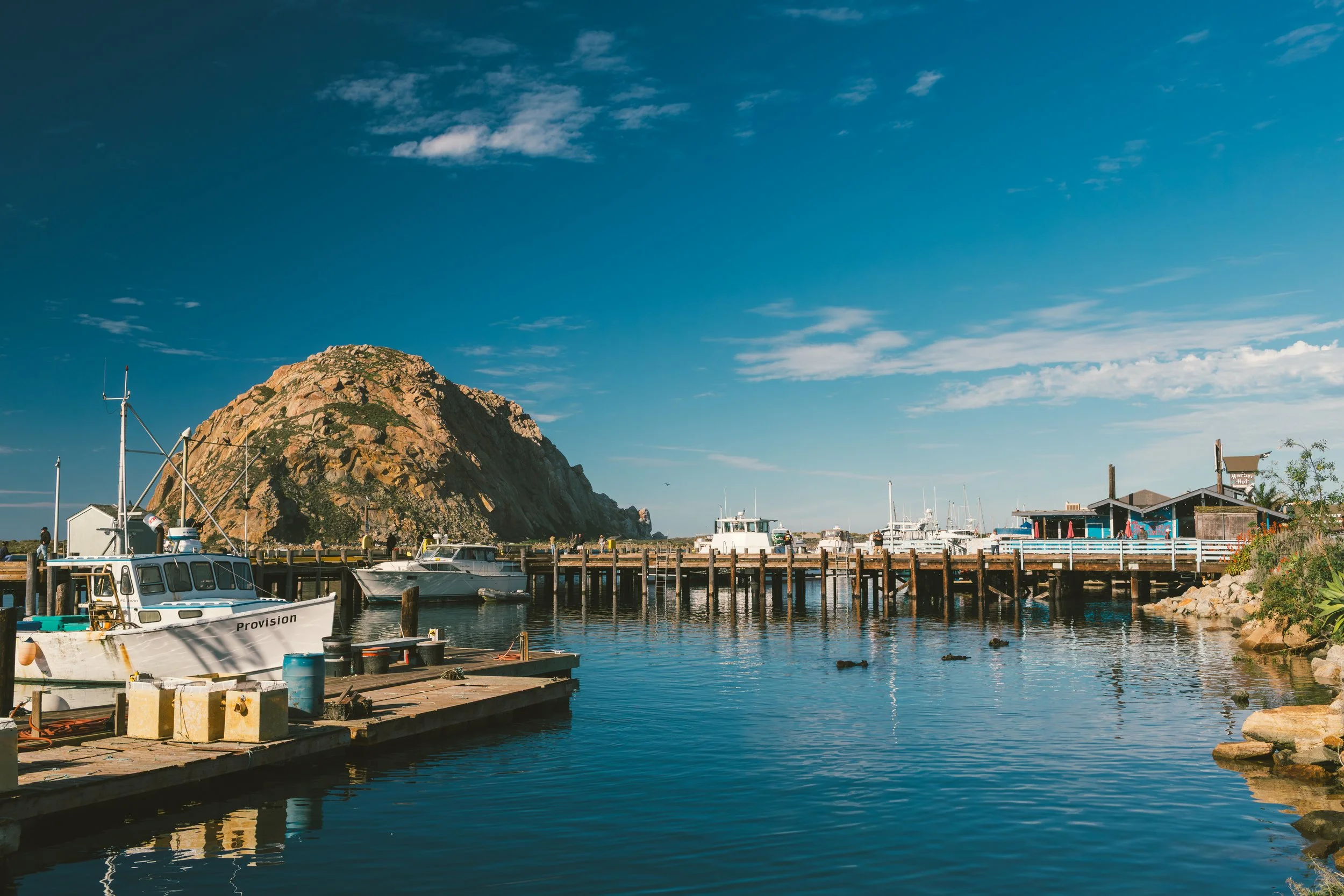 Boats docked at a marina in front of a large rocky hill with a clear blue sky