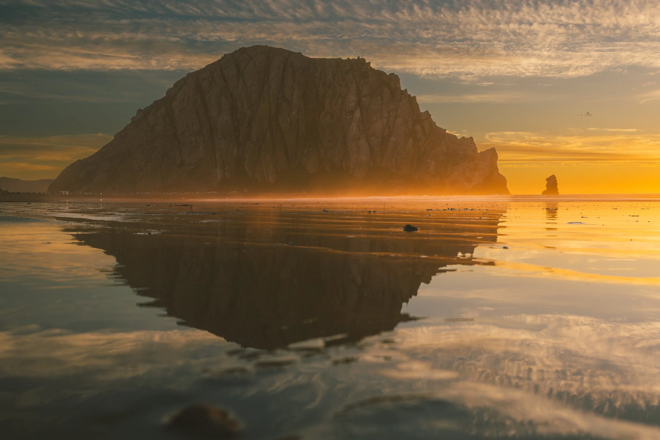 A large rocky cliff is visible at sunset with the sky filled with clouds. The beach below has wet sand reflecting the sky and the cliff, creating a mirror-like effect.