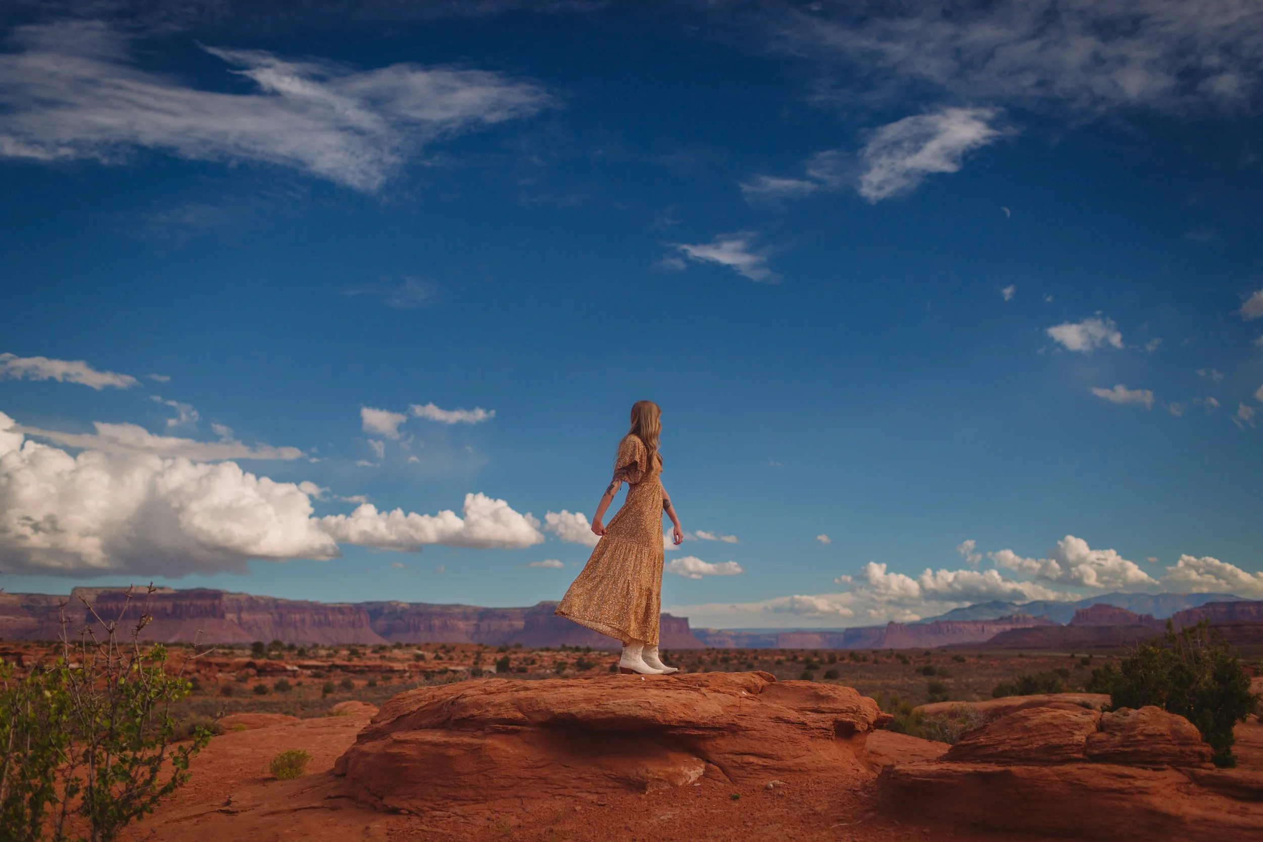 A woman wearing a long dress and white boots stands on red rock in a desert landscape with clouds and mountains in the background.