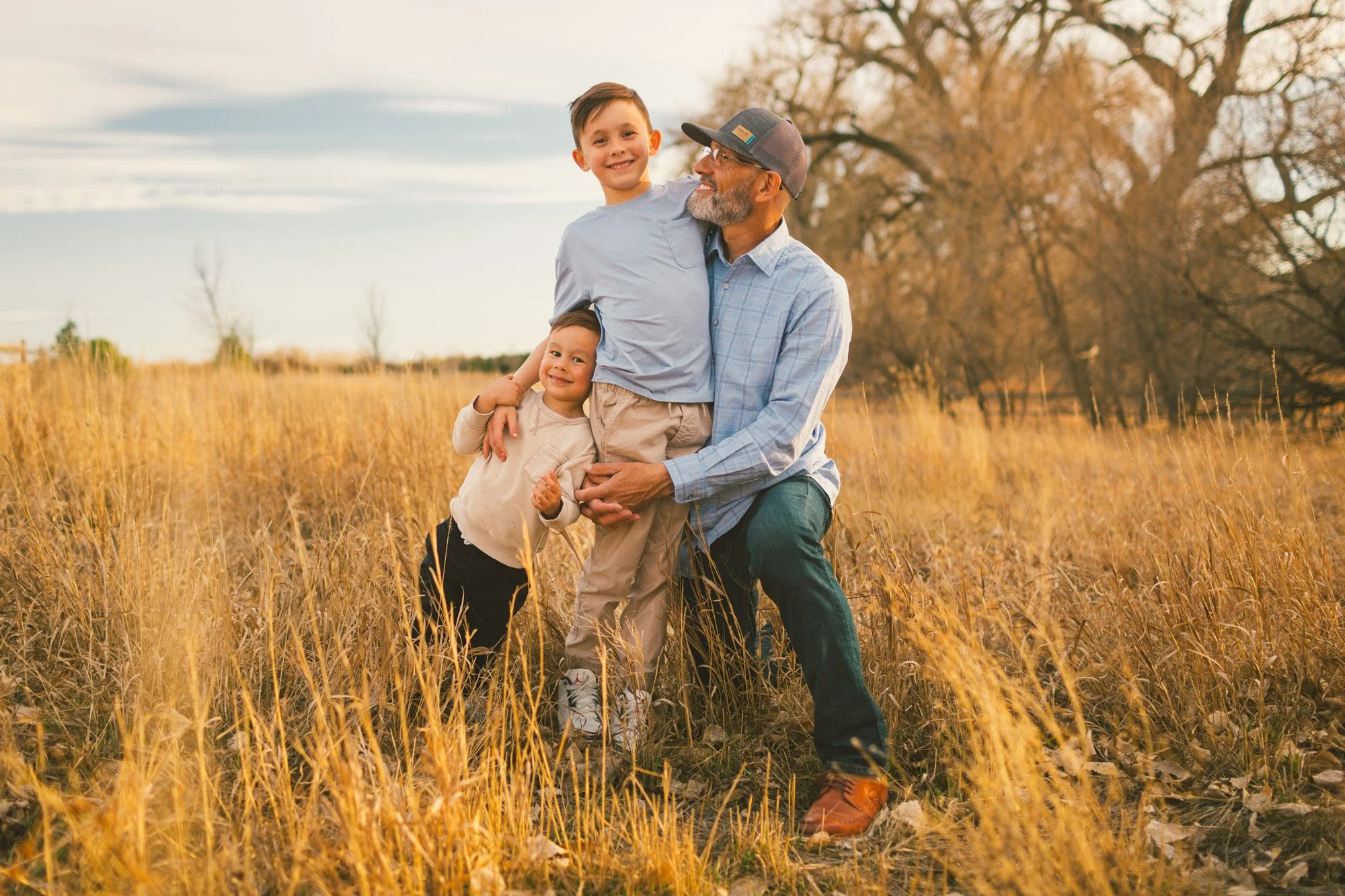A happy grandfather with a gray beard and glasses holding two young boys in a golden field during sunset, with leafless trees in the background.