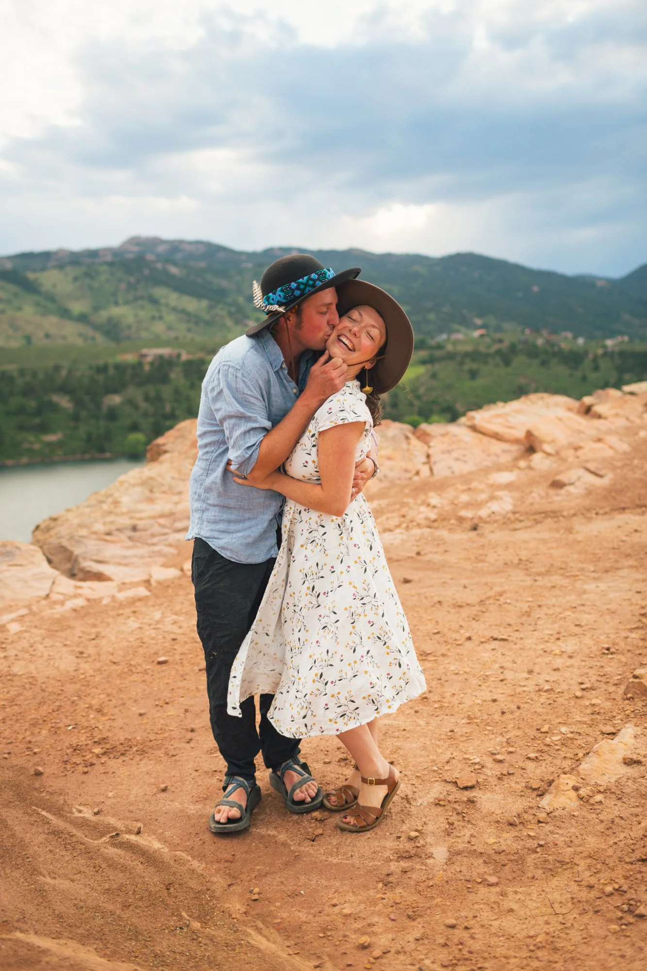 A couple hugging and kissing outdoors on a rocky, reddish terrain with green mountains and cloudy sky in the background.