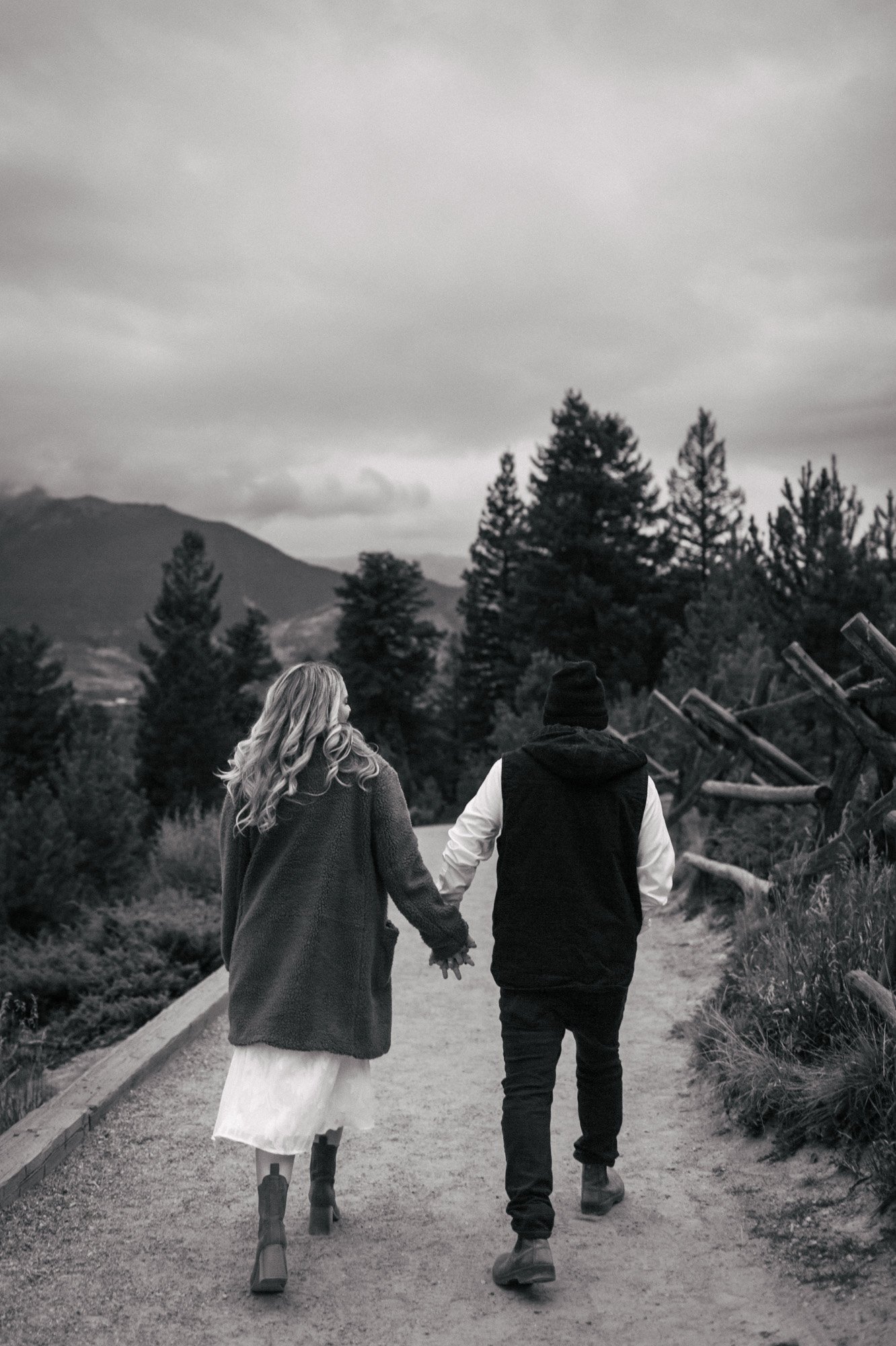 A black-and-white photo of a couple walking hand-in-hand along a tree-lined path in a mountainous area. The woman has long, curly hair and is wearing a coat, skirt, and boots. The man is wearing a beanie, jacket, and pants. The background features tr
