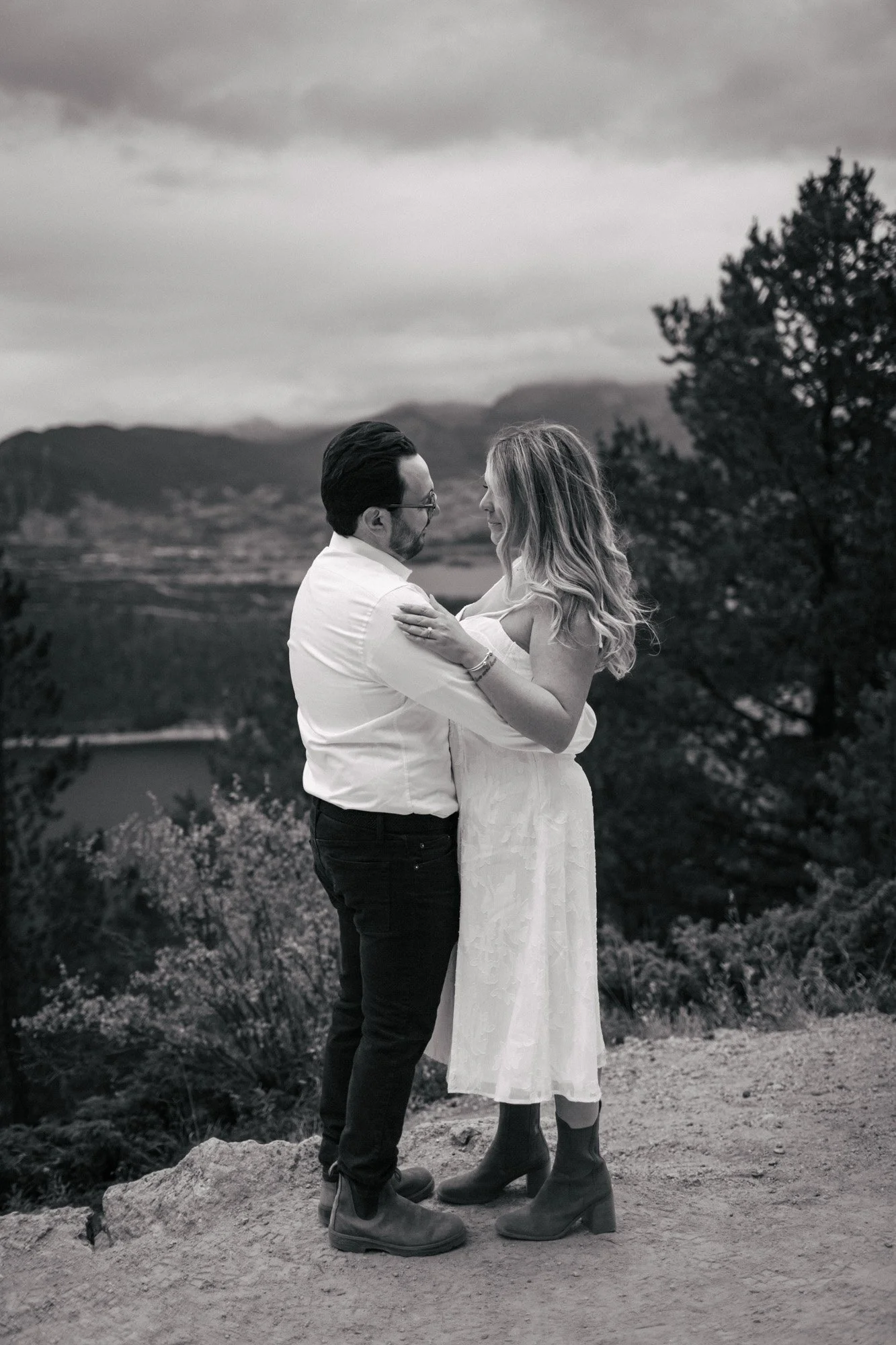 A couple stands together outdoors in a natural landscape with mountains in the background. The man, wearing glasses, a white shirt, and black pants, is holding the woman, who is dressed in a light-colored dress and boots. They are smiling and looking