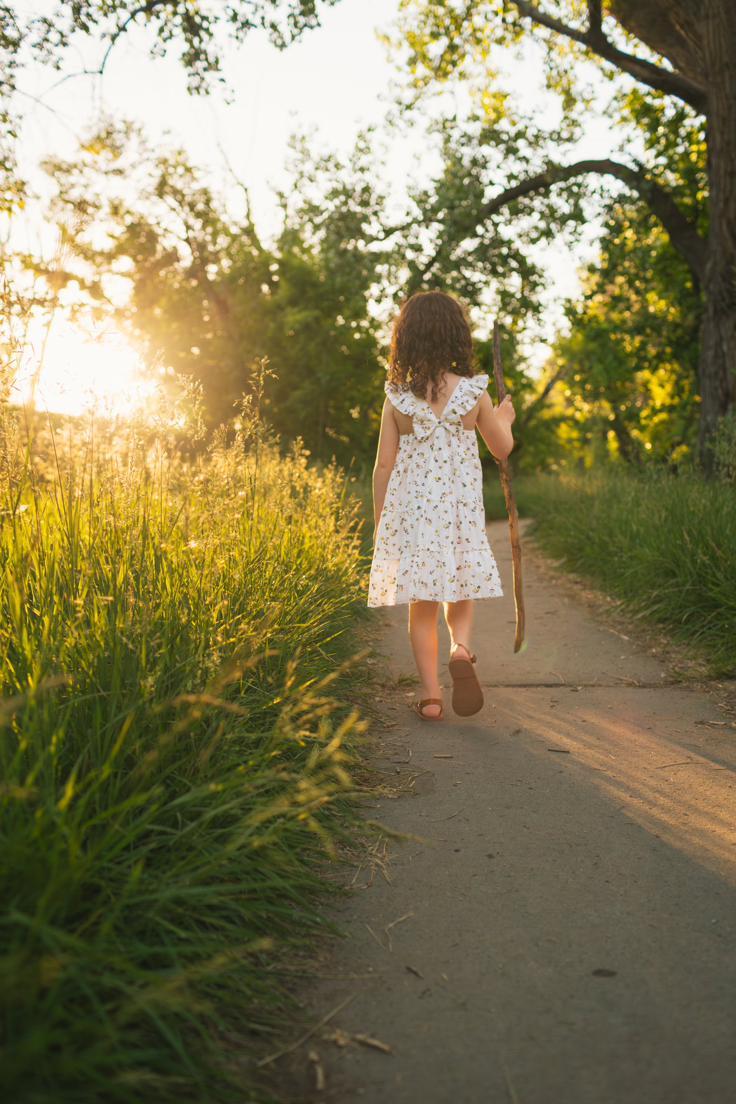 A young girl with curly hair walking along a dirt path during sunset, carrying a stick in her right hand, surrounded by greenery and trees.