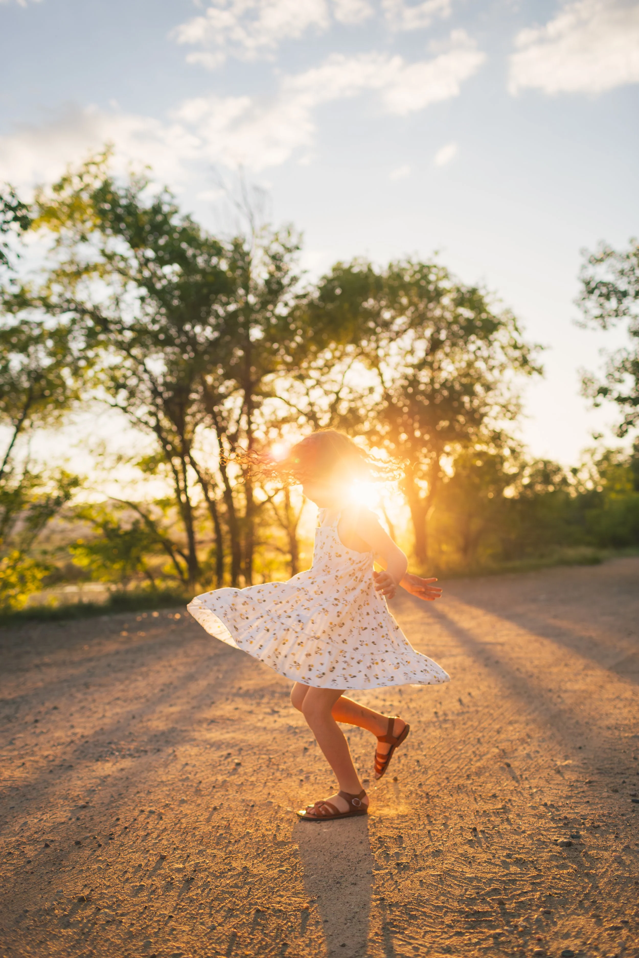 A young girl dancing outdoors on a dirt path during sunset, wearing a white dress with yellow floral patterns, with trees and the setting sun in the background.