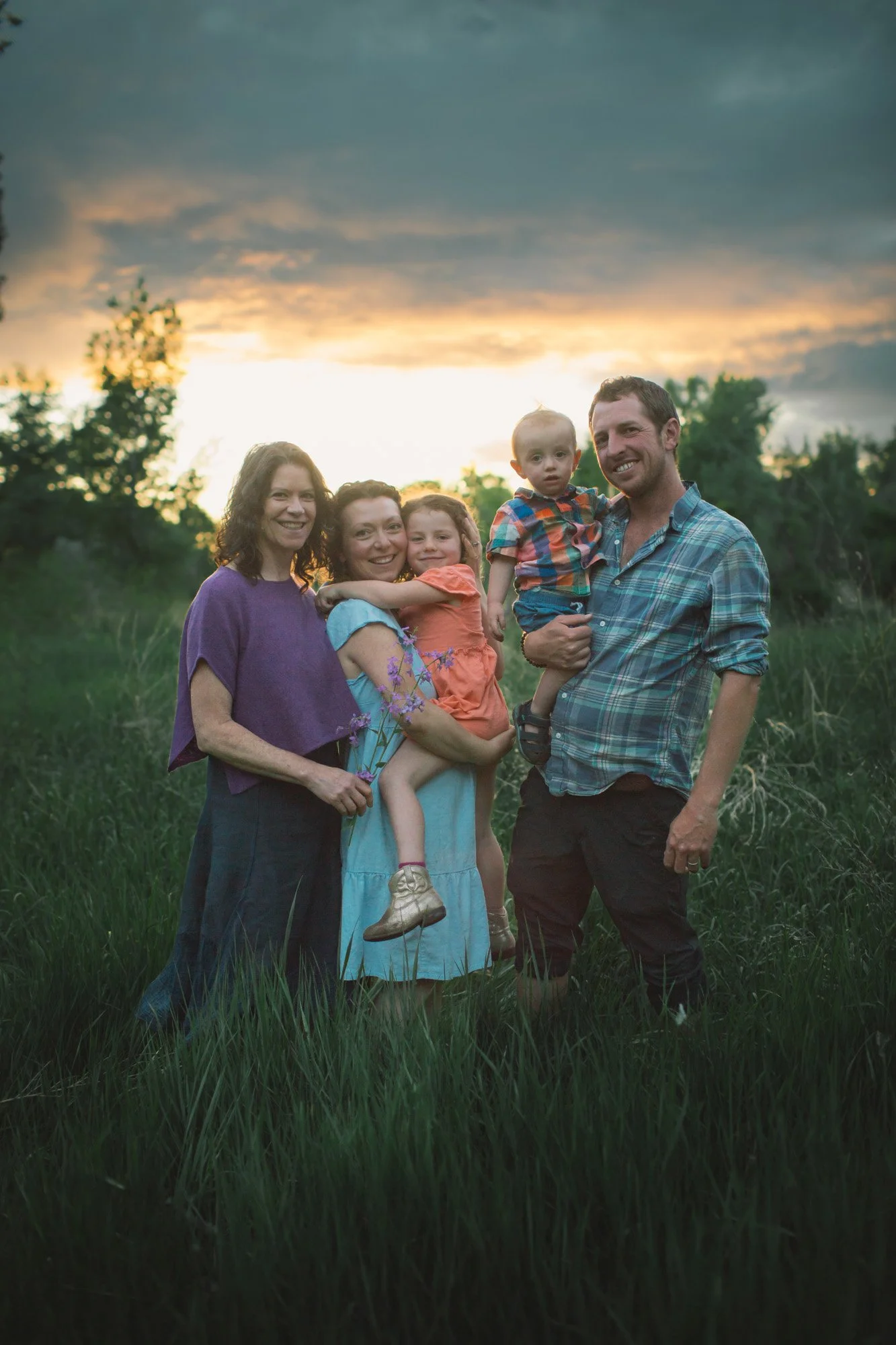A family of five, including two adult women, one adult man, and two children, standing in a grassy field during sunset. The man and one of the women are smiling, and the children are held by the women. The sky is partly cloudy with a dramatic sunset 