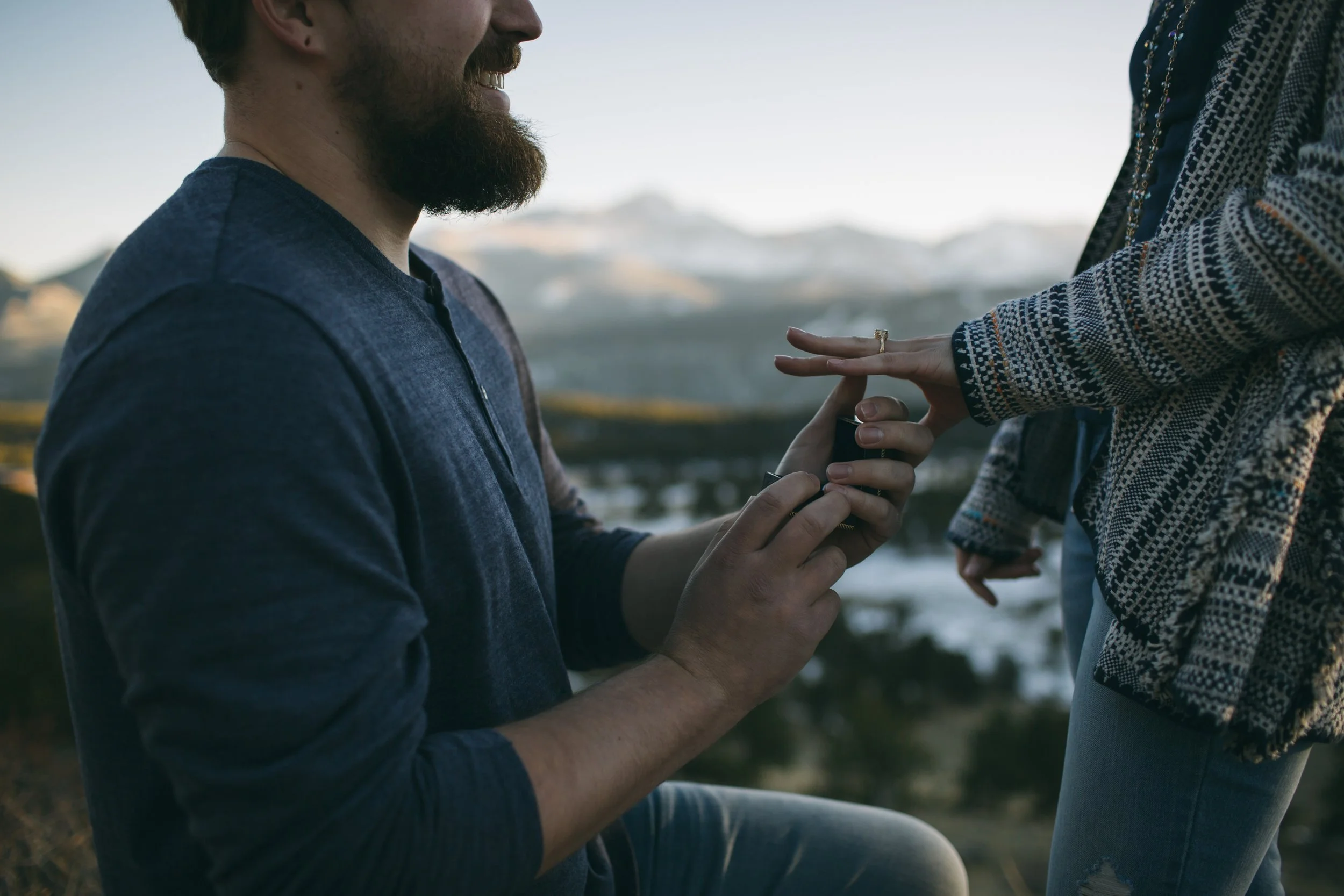 A man is kneeling and placing a ring on a woman's finger during an outdoor proposal at sunset with mountains in the background.