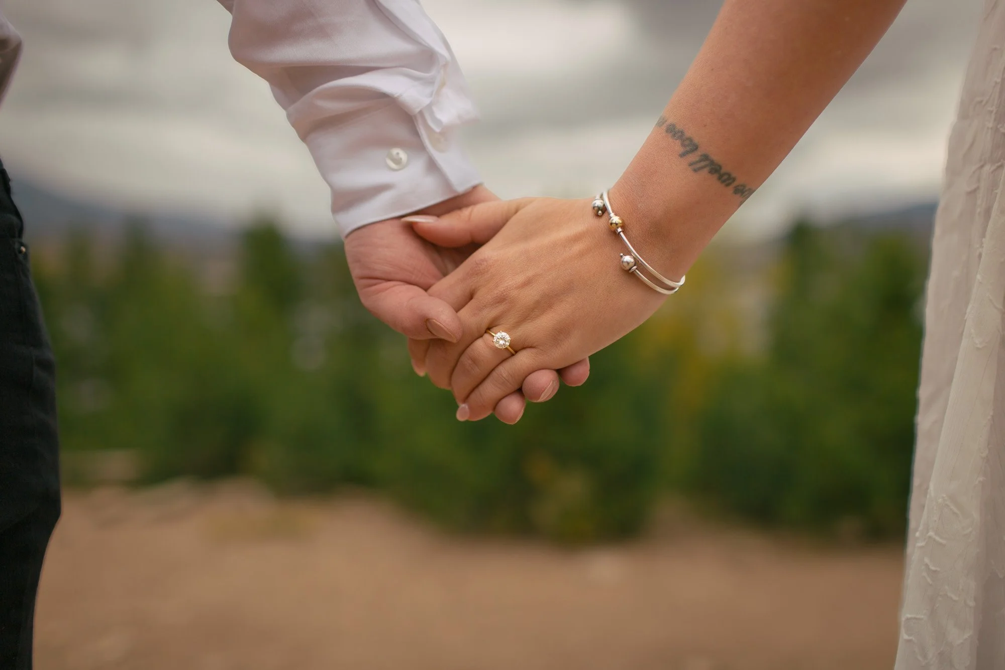 Close-up of two people holding hands, one wearing a white shirt and jewelry including a ring and bracelets, the other dressed in a light-colored outfit, outdoors with a blurred natural background.