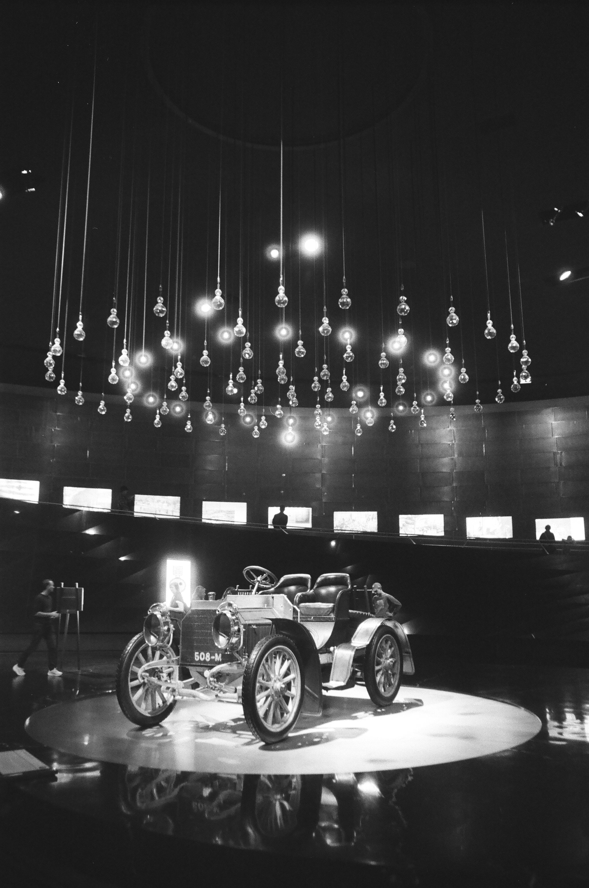 Black and white photograph of a vintage car displayed on a circular platform inside a museum or exhibit space, with people observing and a large chandelier hanging from the ceiling overhead.